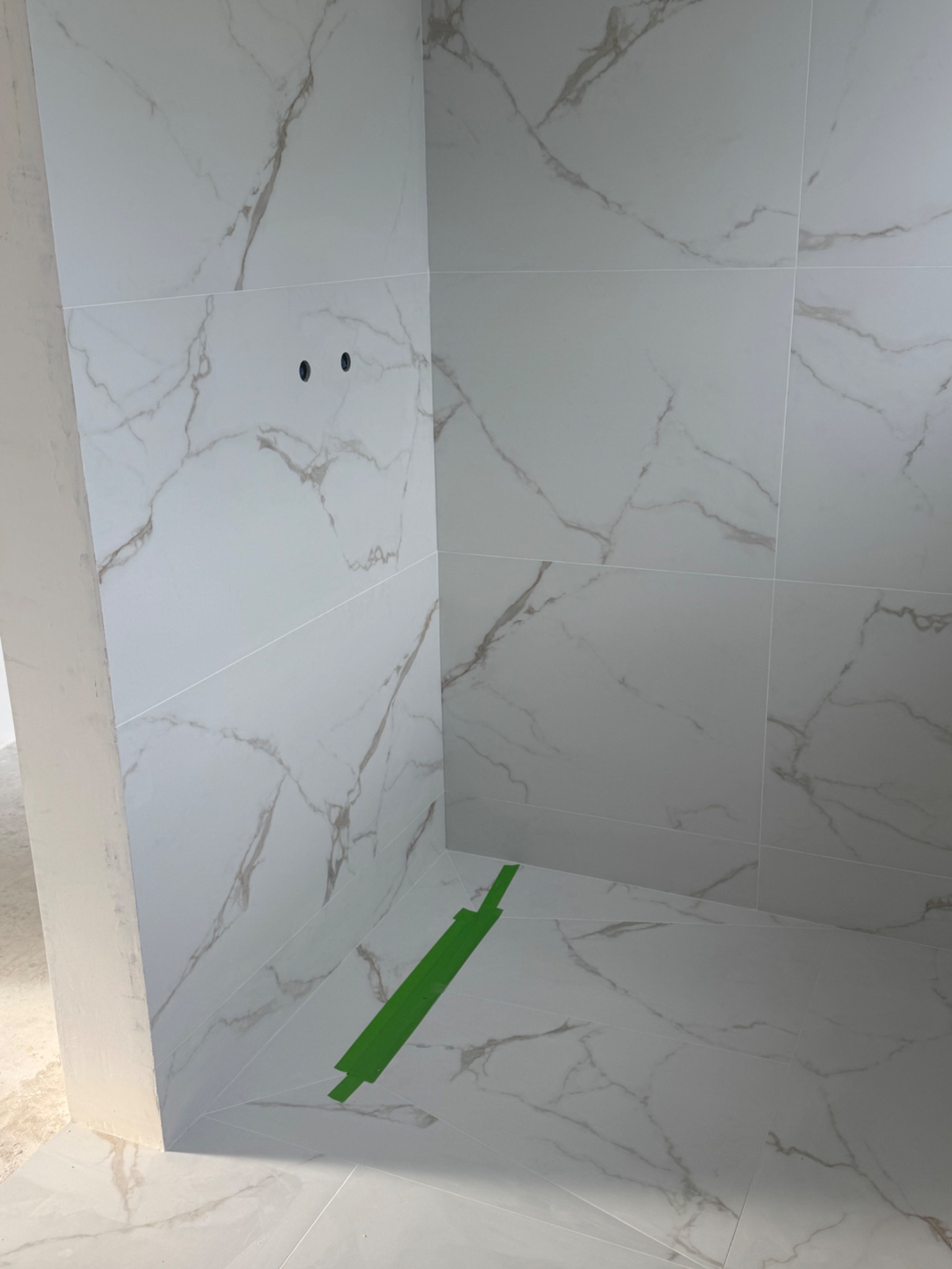 Wykończona kabina prysznicowa z białych płytek imitujących marmur, zabezpieczona taśmą malarską. Widoczne otwory na armaturę.