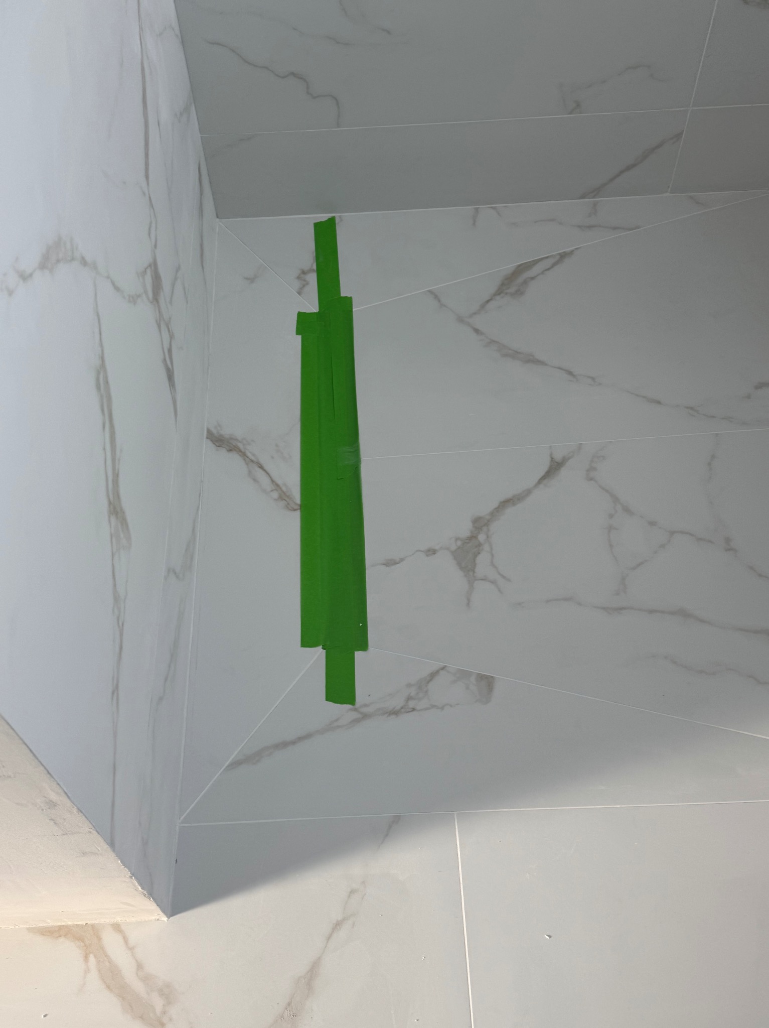 Wykończona ściana i podłoga w łazience z białych, marmurkowych płytek, zabezpieczona zieloną taśmą malarską w rogu. Widok z dołu.