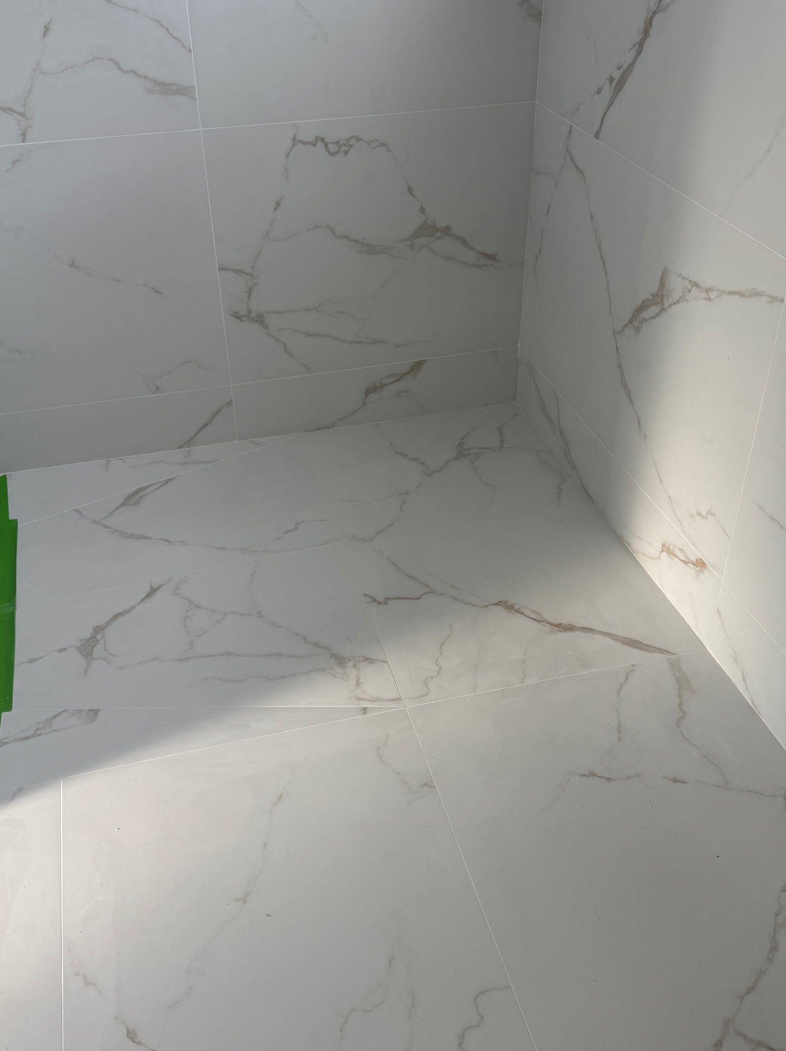 Wykończona podłoga i ściany w łazience wyłożone białymi płytkami imitującymi marmur z delikatnymi, szarymi żyłkami. W rogu widoczny fragment zielonej taśmy malarskiej.