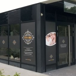 Druknij.to - Elewacja salonu 'Tivore' z naklejkami na oknach prezentującymi logo, grafikę oka z rzęsami i detale oferty, utrzymana w czarno-białej kolorystyce z elementami złota.
