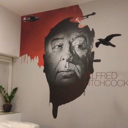 Druknij.to - Malowidło ścienne z portretem Alfreda Hitchcocka i motywem ptaków, wykonane w czerni i czerwieni. Obok stoi roślina doniczkowa, a w tle widać fragment mebla.