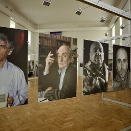 Druknij.to - Wystawa portretów w jasnym wnętrzu. Fotografie wiszą na linkach, ukazując wizerunki osób. Parkiet odbija światło, a ekspozycja jest minimalistyczna i elegancka.