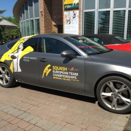 Druknij.to - Szary samochód Audi z grafiką Squash Kahuna European Team Championships, Wojtek Nowisz Polska, zaparkowany na brukowanym chodniku, z budynkiem w tle.
