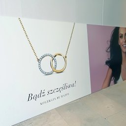 Druknij.to - Druk wielkoformatowy: Baner reklamowy z wizerunkiem kobiety i biżuterii, hasło 'Bądź szczęśliwa! Kolekcja BE HAPPY' na białym tle, ustawiony na podłodze.