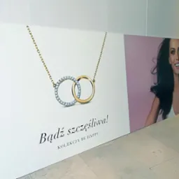 Druk wielkoformatowy: Baner reklamowy z wizerunkiem kobiety i biżuterii, hasło 'Bądź szczęśliwa! Kolekcja BE HAPPY' na białym tle, ustawiony na podłodze.