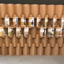 Ekspozycja ulotek na tekturowym stojaku z rośliną w tle. Ulotki prezentują grafiki i teksty o tematyce ekologicznej i zrównoważonego rozwoju.