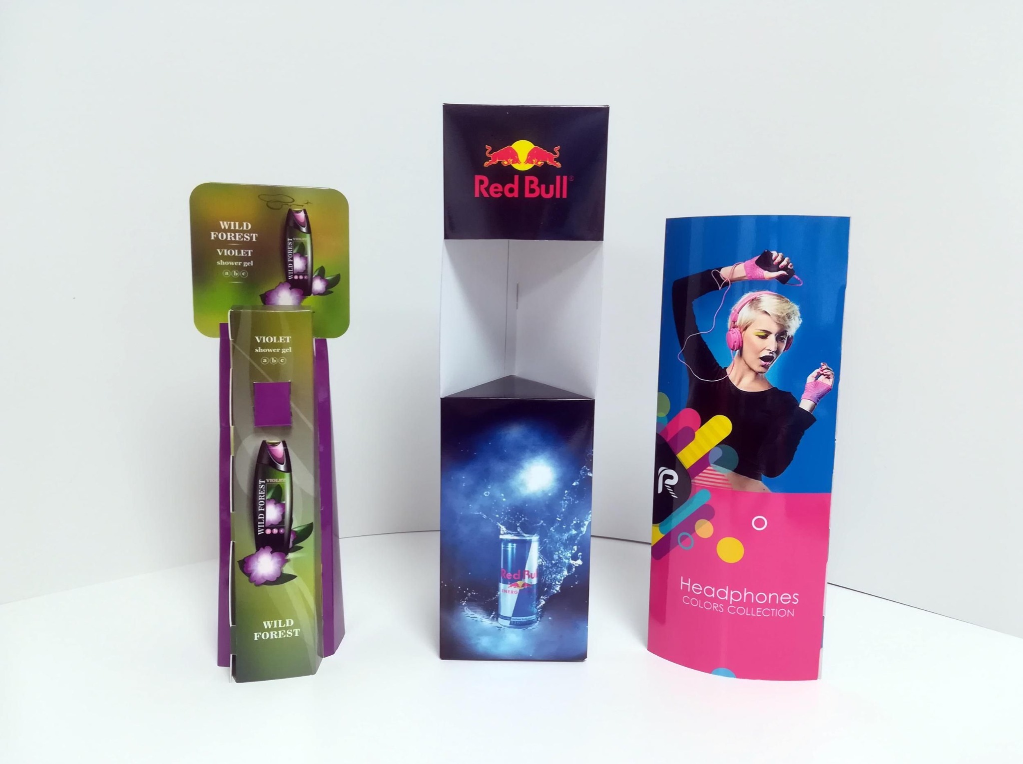 Ekspozytory reklamowe: kartonowy stojak na kosmetyki Wild Forest, trójkątny display Red Bull i cylinder z grafiką słuchawek. Białe tło, studyjne oświetlenie.
