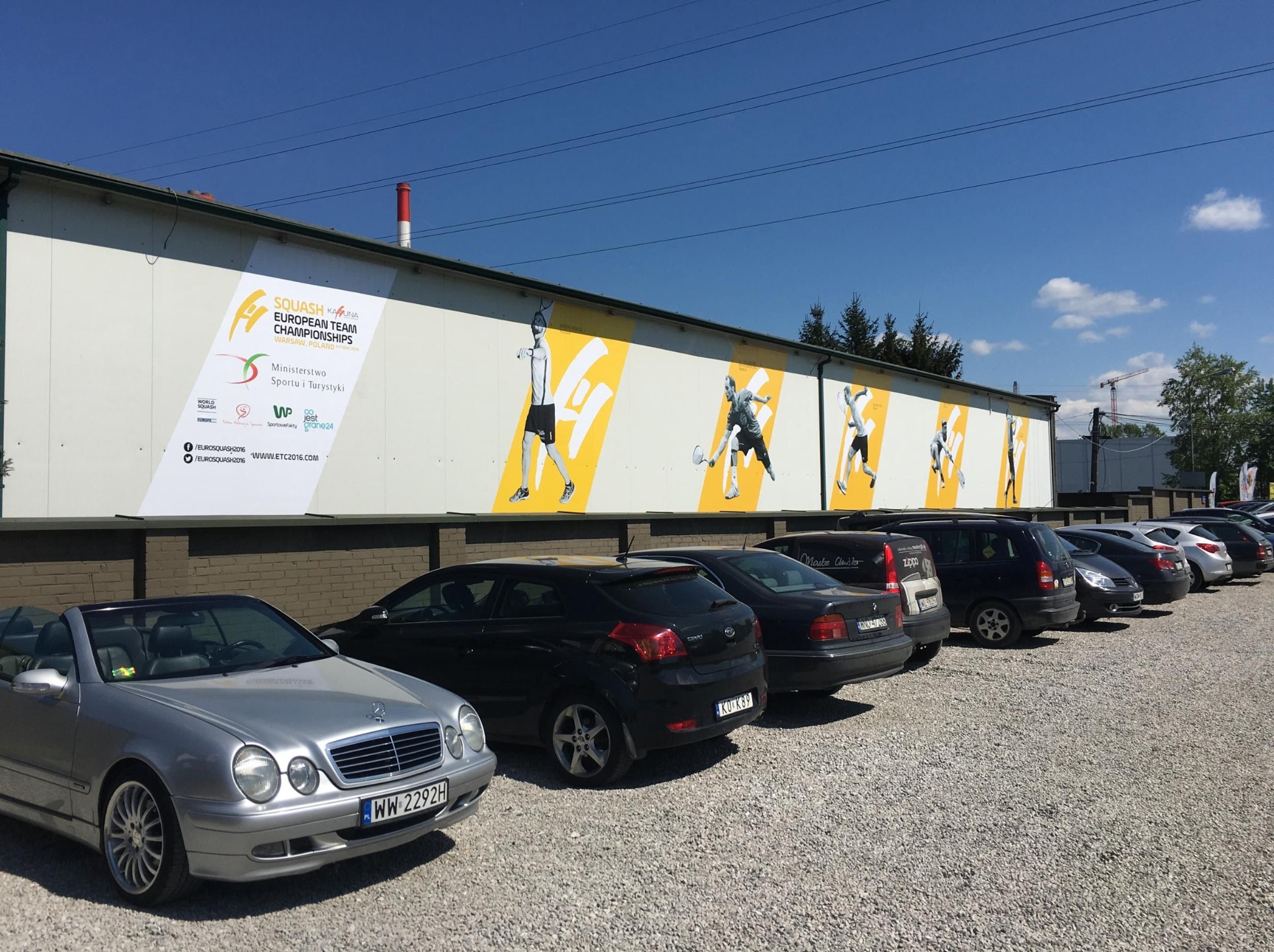 Druk wielkoformatowy: Billboard reklamowy zawodów squash na ścianie budynku, zaparkowane samochody w pierwszym planie, błękitne niebo w tle.
