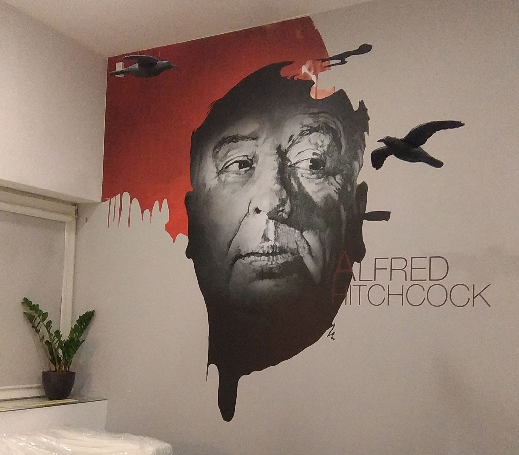 Malowidło ścienne z portretem Alfreda Hitchcocka i motywem ptaków, wykonane w czerni i czerwieni. Obok stoi roślina doniczkowa, a w tle widać fragment mebla.