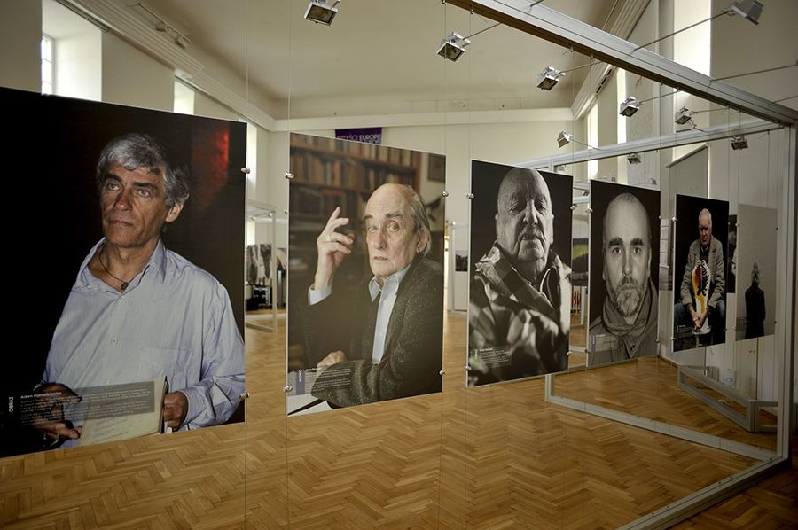 Wystawa portretów w jasnym wnętrzu. Fotografie wiszą na linkach, ukazując wizerunki osób. Parkiet odbija światło, a ekspozycja jest minimalistyczna i elegancka.