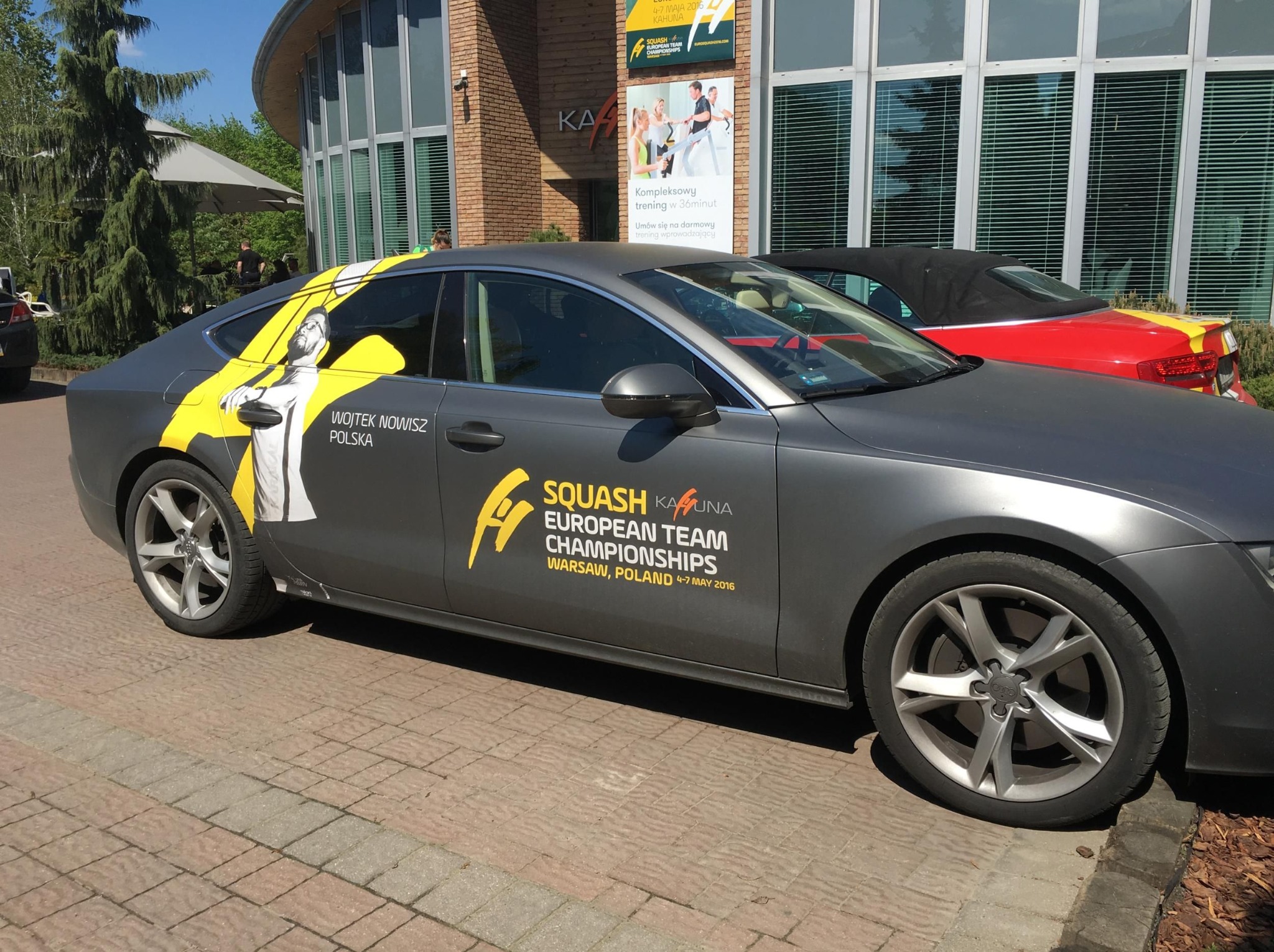 Szary samochód Audi z grafiką Squash Kahuna European Team Championships, Wojtek Nowisz Polska, zaparkowany na brukowanym chodniku, z budynkiem w tle.