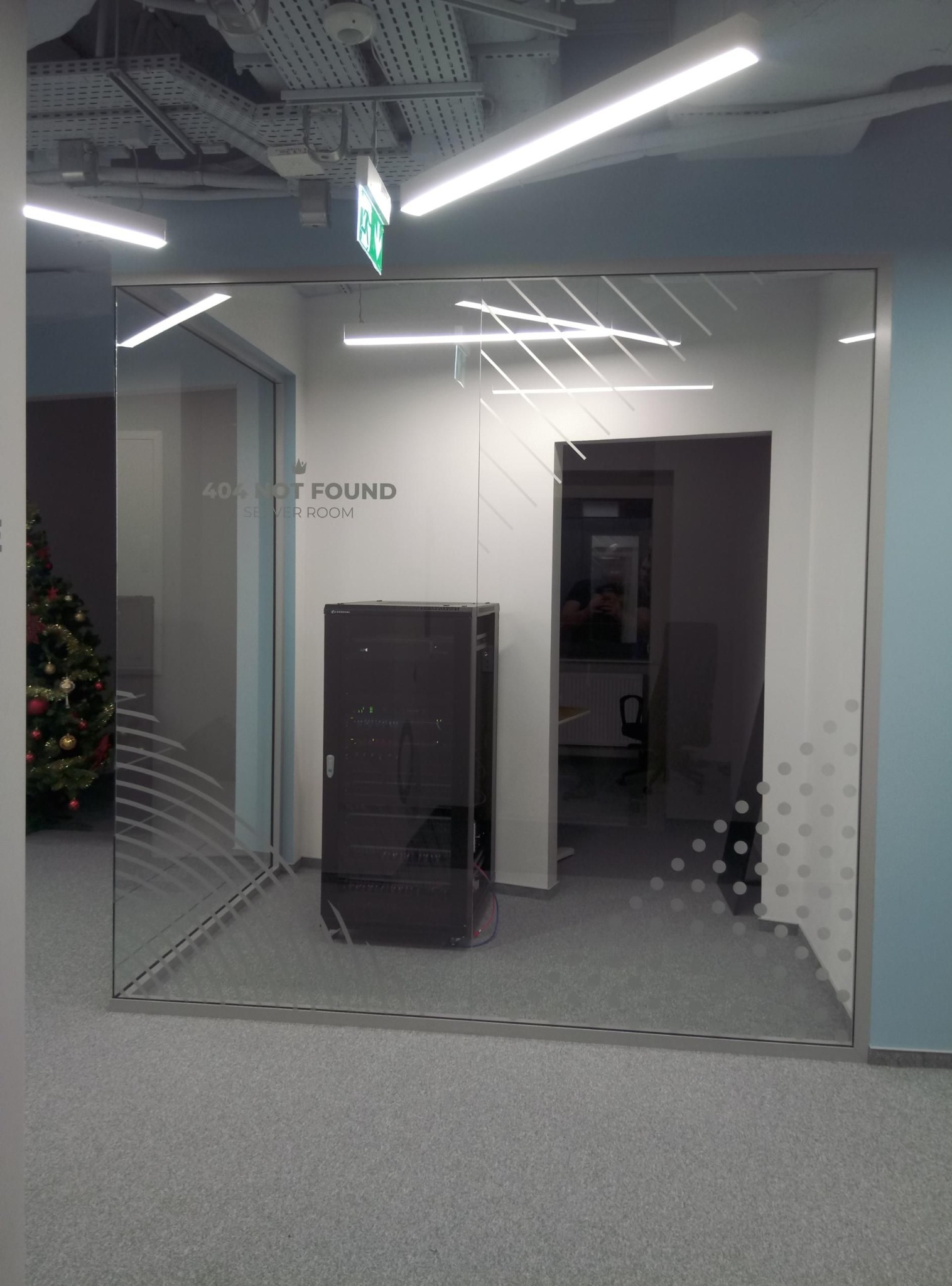 Szklana ściana z napisem '404 NOT FOUND SERVER ROOM' w nowoczesnym biurze. W tle serwerownia i choinka, widoczne elementy instalacji pod sufitem.