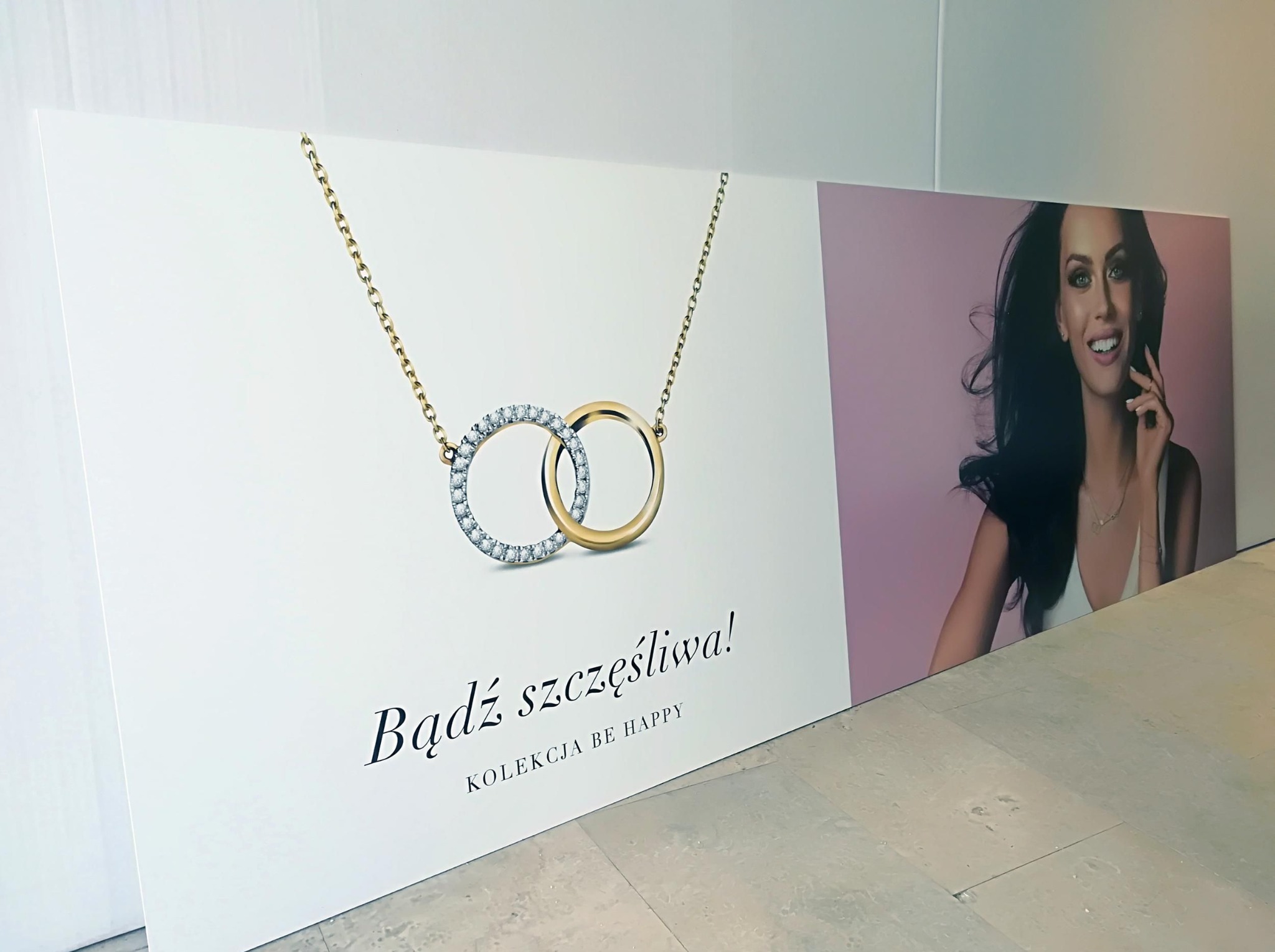 Druk wielkoformatowy: Baner reklamowy z wizerunkiem kobiety i biżuterii, hasło 'Bądź szczęśliwa! Kolekcja BE HAPPY' na białym tle, ustawiony na podłodze.