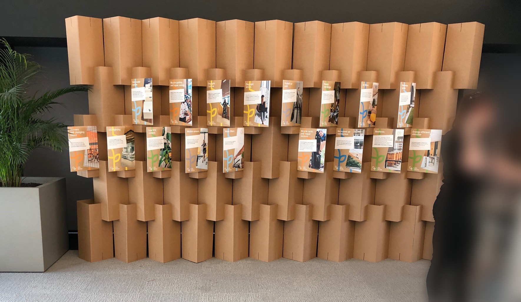 Ekspozycja ulotek na tekturowym stojaku z rośliną w tle. Ulotki prezentują grafiki i teksty o tematyce ekologicznej i zrównoważonego rozwoju.