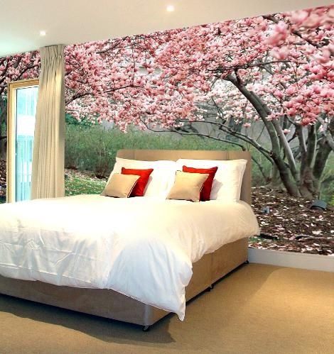 Sypialnia z łóżkiem na tle fototapety z kwitnącą magnolią. Naturalne światło z okna po lewej stronie. Beżowa podłoga i rama łóżka.