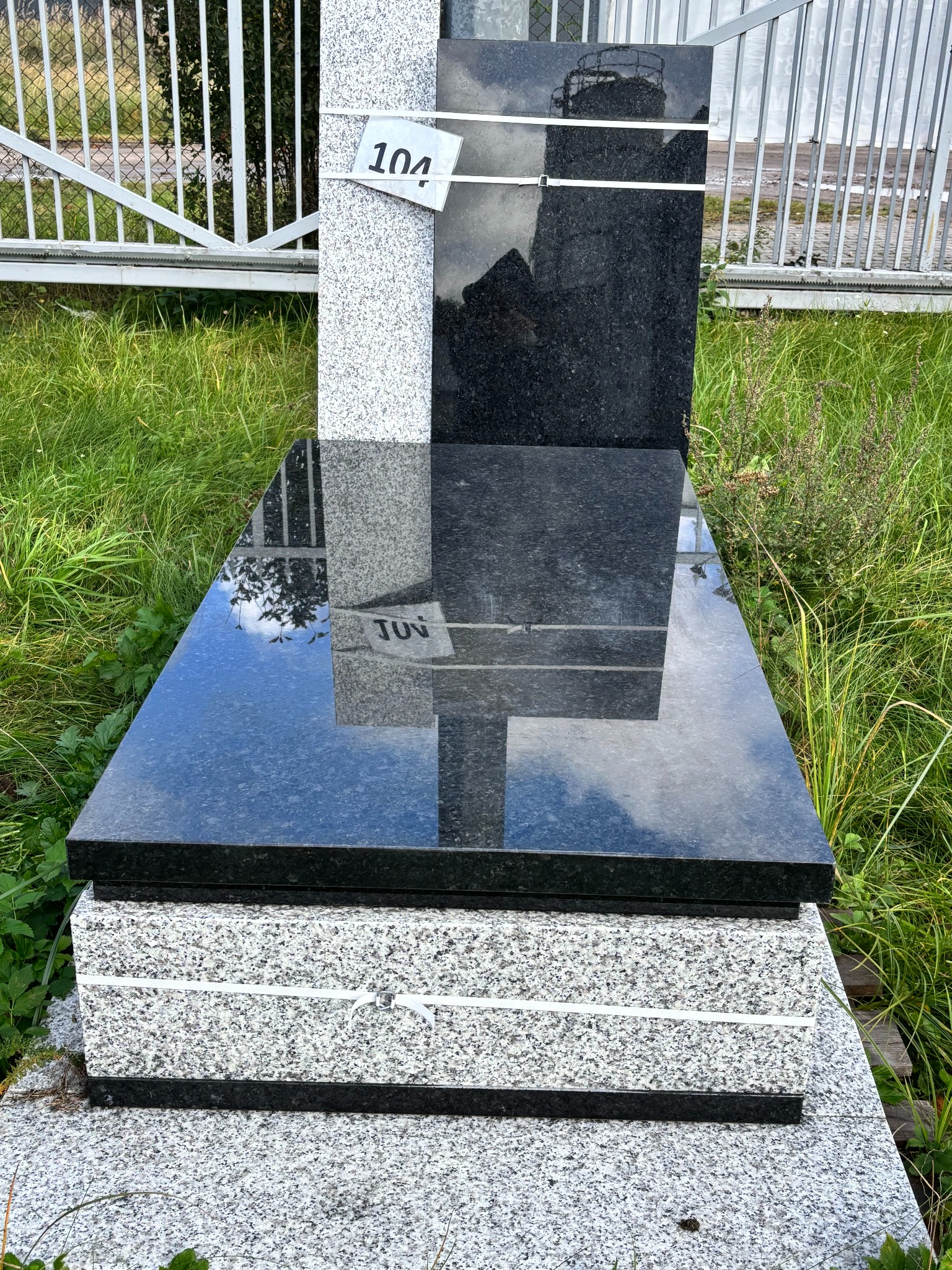 Granitowy pomnik w odcieniach szarości i czerni, z widoczną tabliczką z numerem '104' i odbiciem nieba na polerowanej powierzchni, otoczony zielenią.