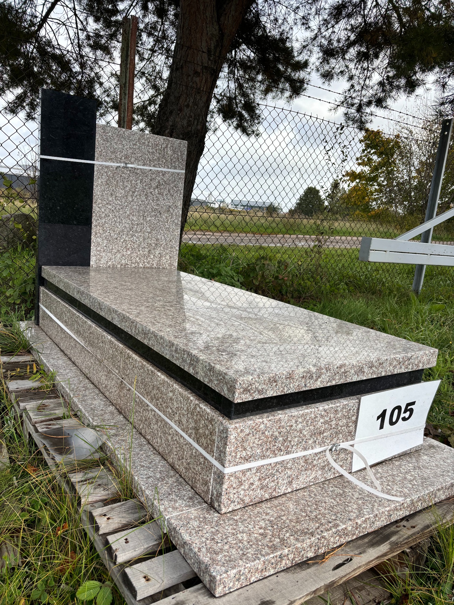 Granitowy nagrobek w odcieniach szarości i czerni, zabezpieczony taśmami, numer 105 na białej etykiecie, na drewnianej palecie, w tle ogrodzenie z siatki.