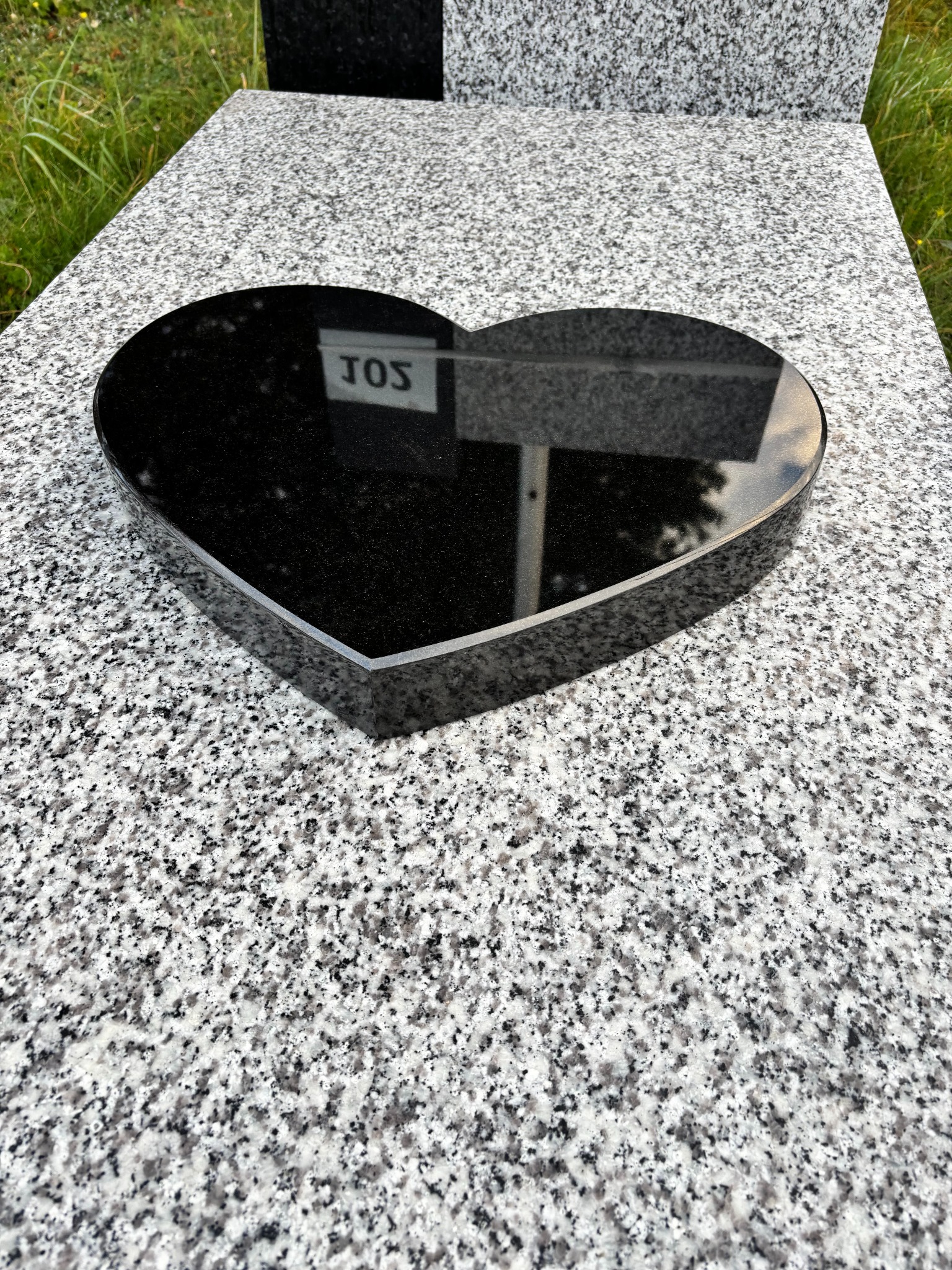 Czarny, granitowy element w kształcie serca na nagrobku z szarego, nakrapianego granitu. Widok z góry, naturalne oświetlenie.