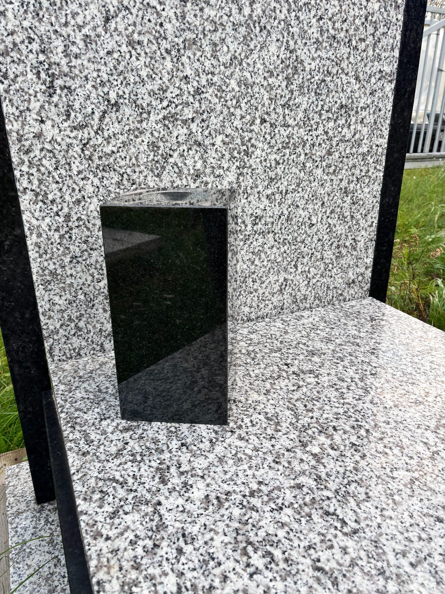 Granitowy wazon w kształcie prostopadłościanu, z polerowanego czarnego kamienia, na tle jasnoszarej płyty nagrobkowej.