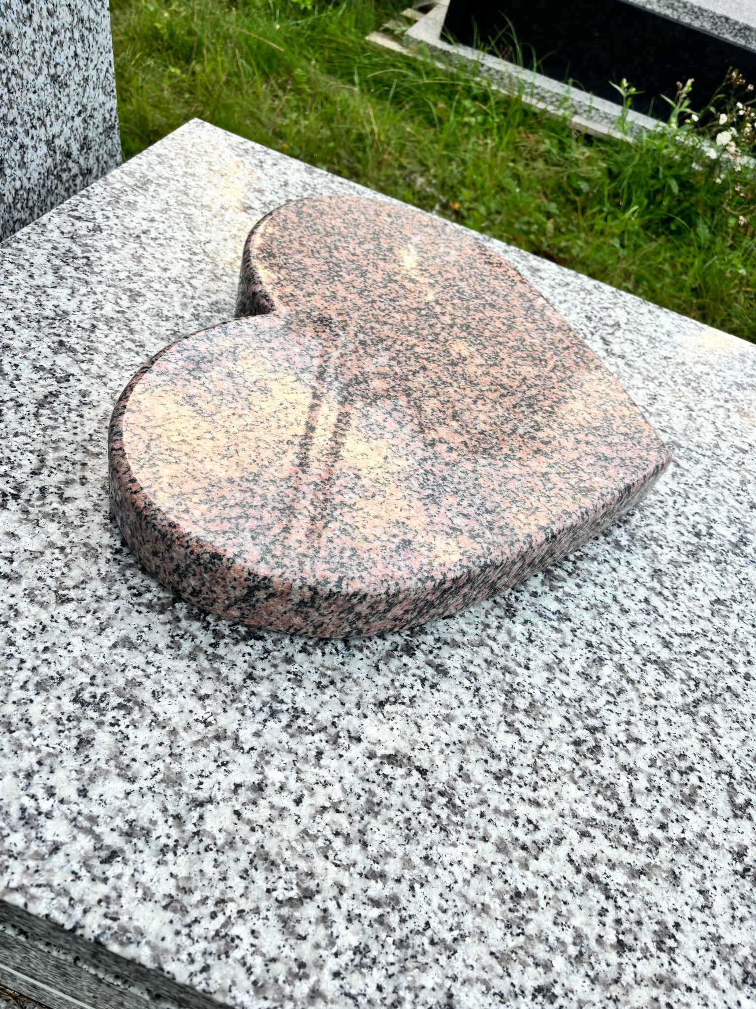Granitowe serce leży na jasnoszarym nagrobku. W tle zieleń trawy. Element dekoracyjny z kamienia w kształcie serca, wykonany z różowego granitu.