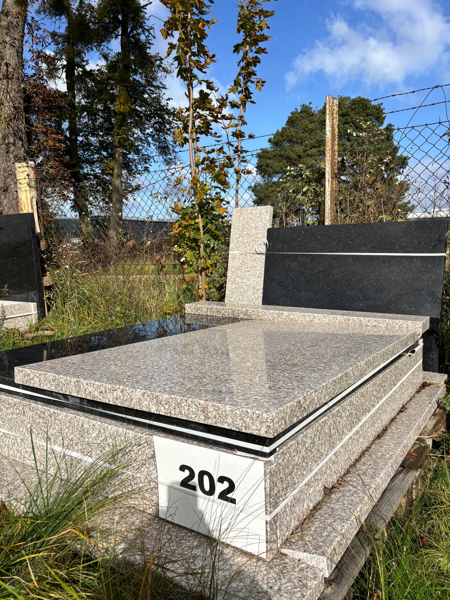 Jasny, granitowy pomnik z numerem '202' w otoczeniu zieleni, z widocznym czarnym nagrobkiem w tle i niebieskim niebem. Prezentacja nagrobka.