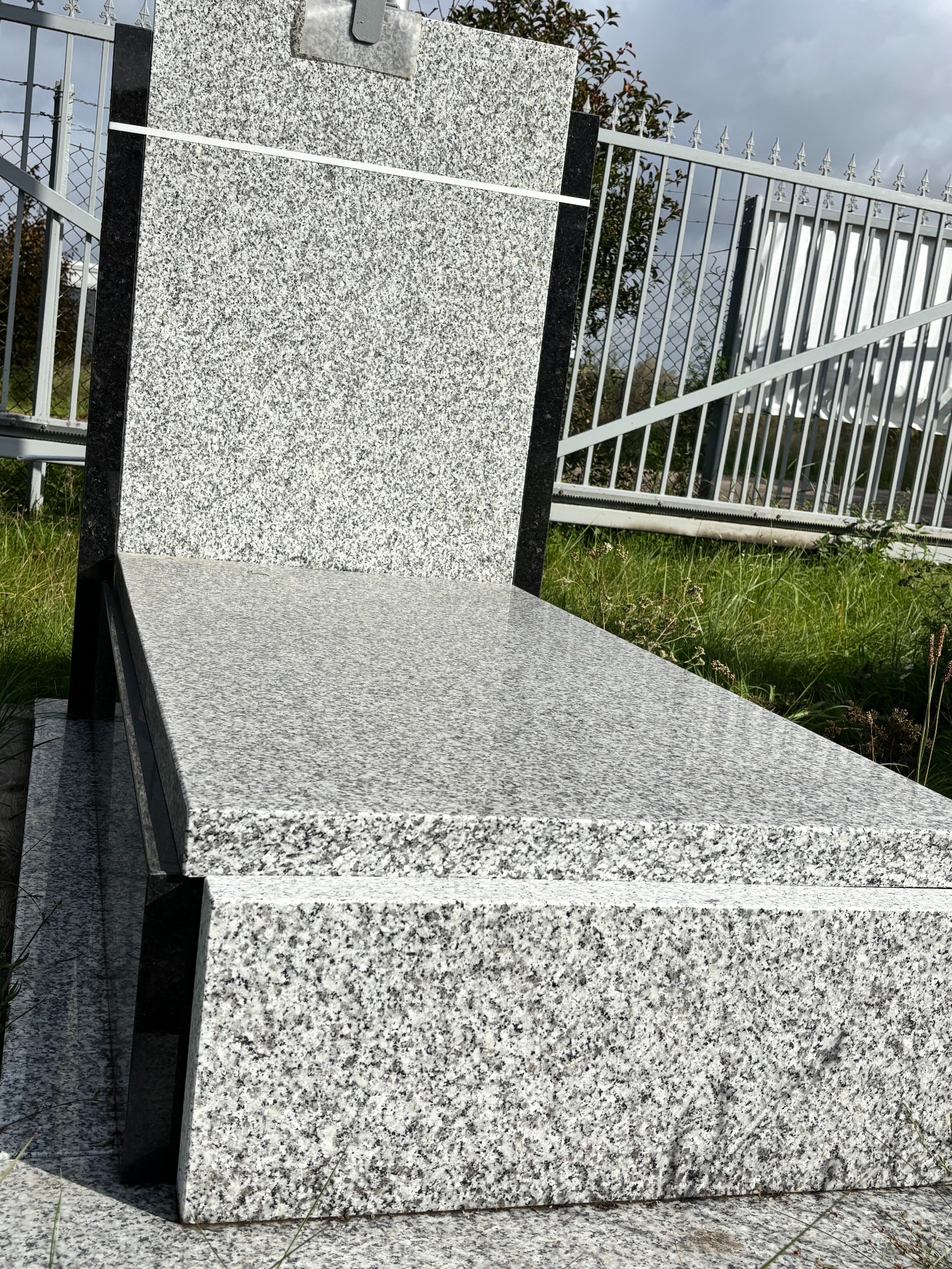 Jasnoszary, granitowy nagrobek z ciemnymi elementami, ustawiony na zewnątrz, na tle metalowego ogrodzenia i zieleni. Widoczne detale faktury kamienia.