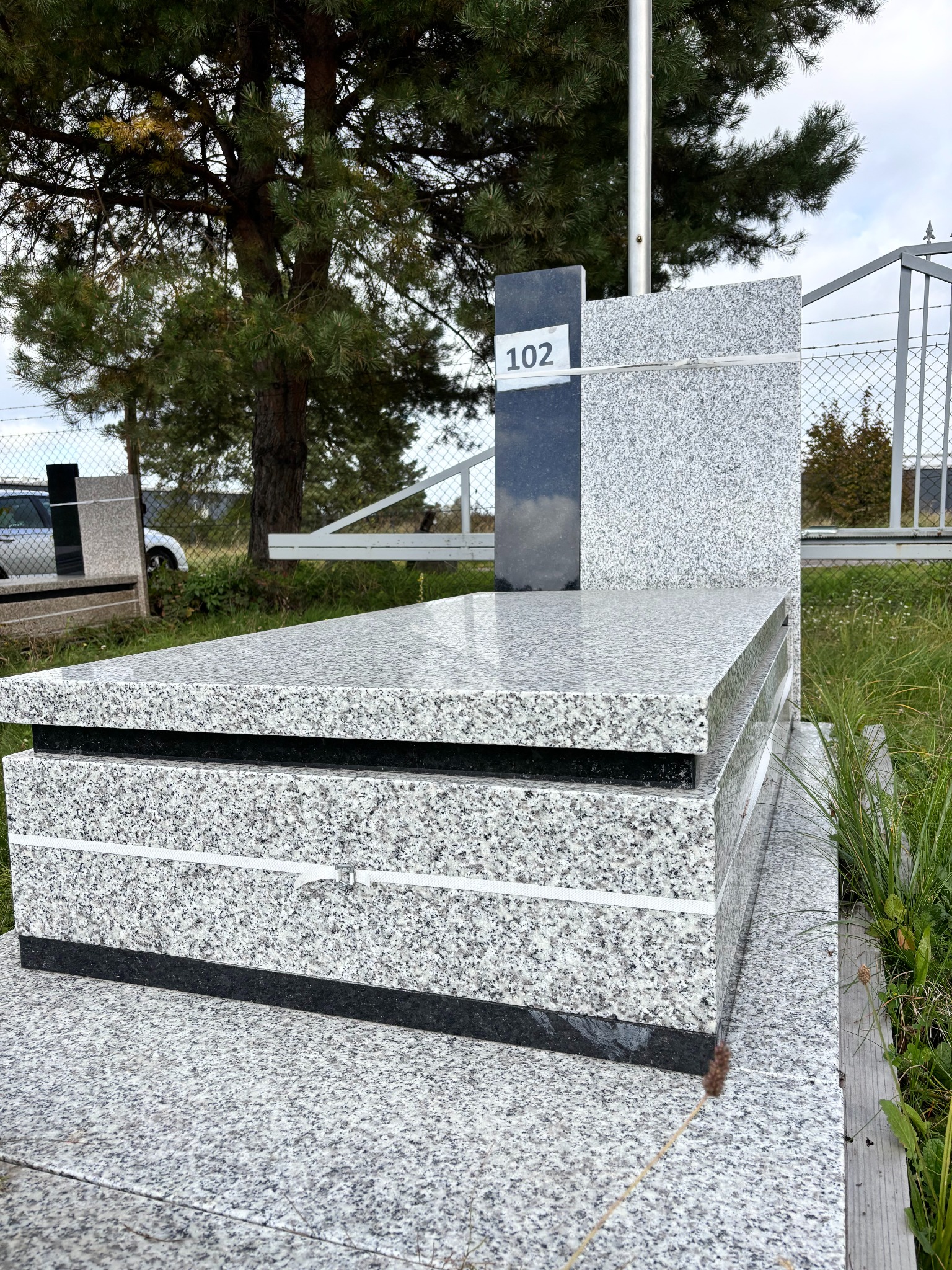 Jasnoszary, granitowy pomnik z czarnym akcentem i numerem '102' na tabliczce, otoczony zielenią i ogrodzeniem z siatki. Widok z boku, w tle drzewo.