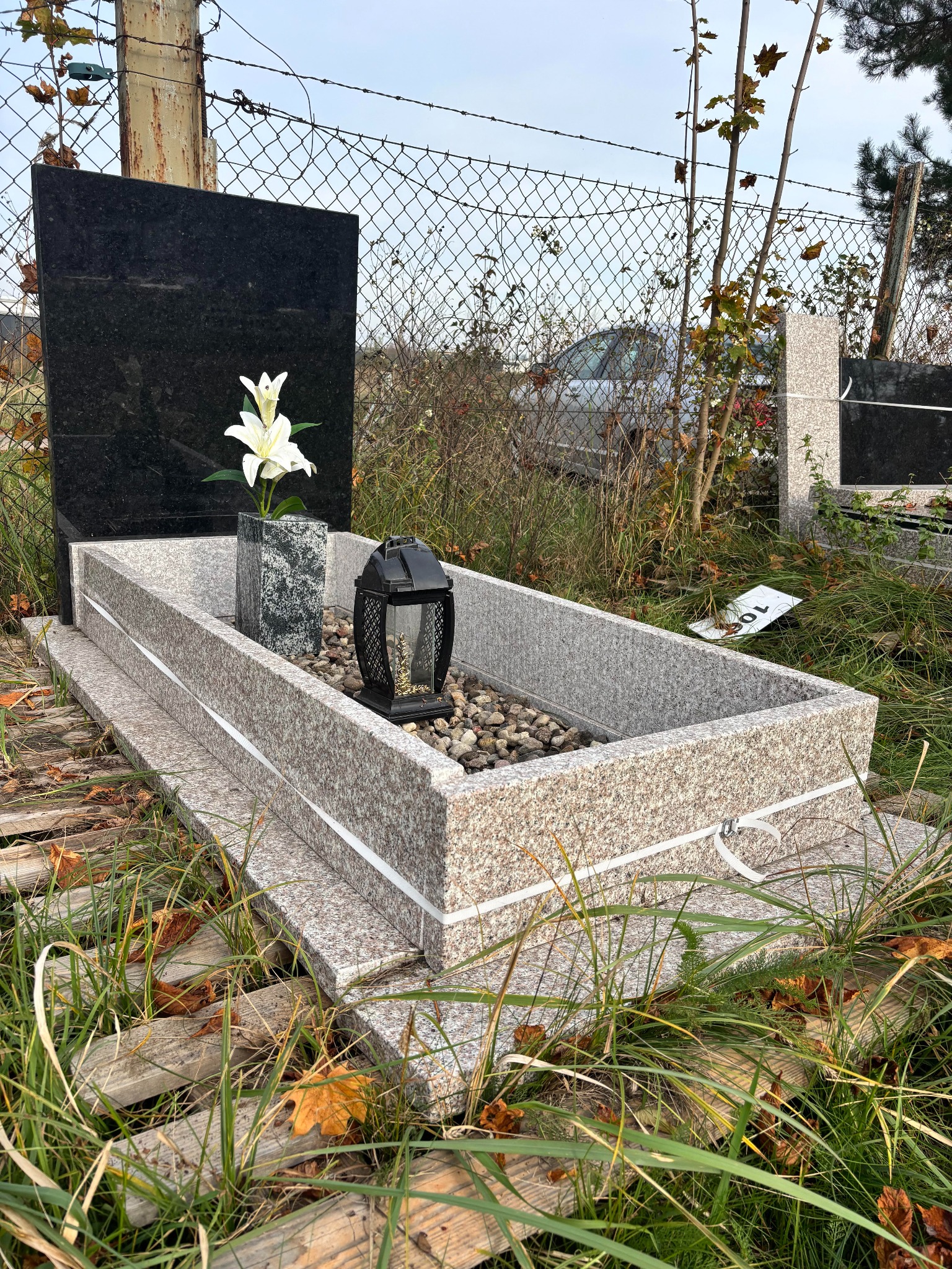 Granitowy pomnik z czarną tablicą, białymi liliami w wazonie i lampionem. Otoczenie: kamyki, trawa, drewniane deski. W tle siatka ogrodzeniowa i fragment innego pomnika.