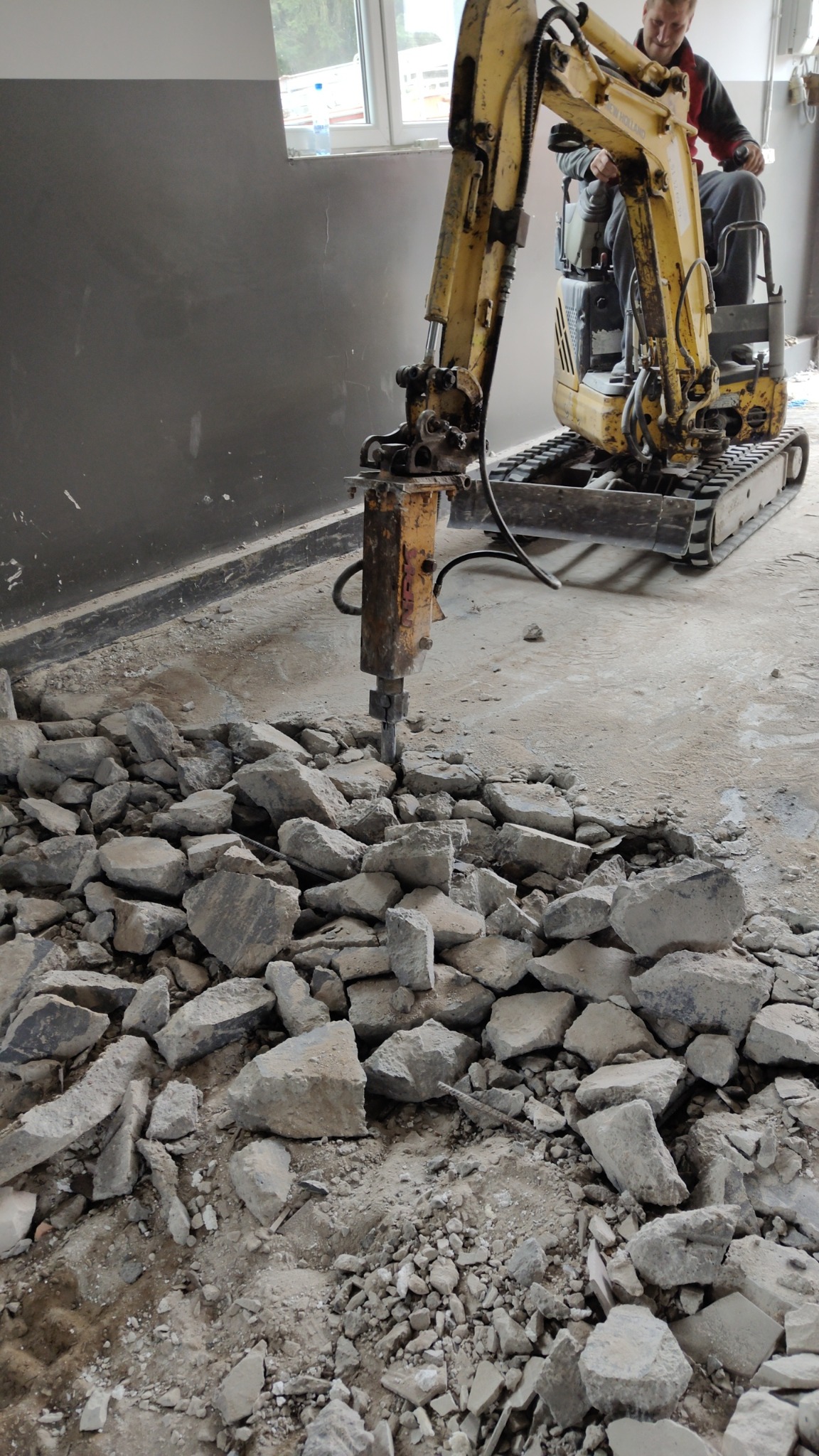 Żółta minikoparka z wiertłem pneumatycznym kruszy betonową posadzkę w szarym pomieszczeniu. Operator w kabinie. Widoczne gruz i pył.