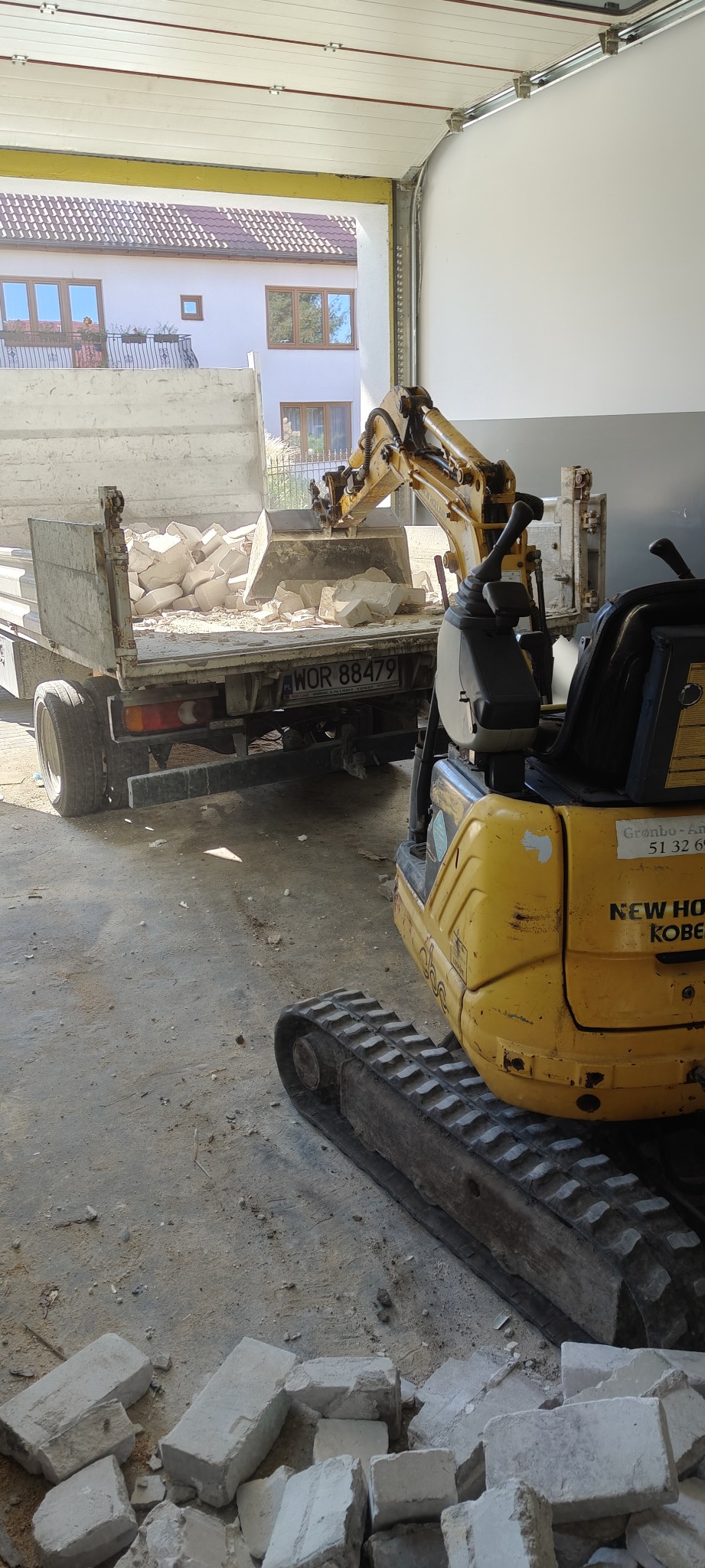Żółta mini koparka załadowuje gruz do skrzyni białej ciężarówki w garażu; widoczny numer rejestracyjny WOR 88479. Część domu widoczna w tle.