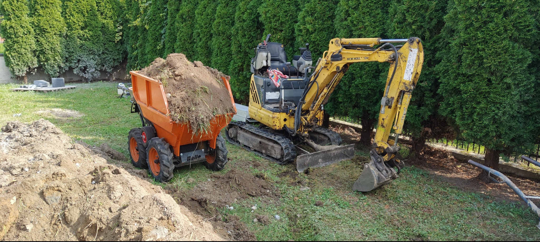 Żółta mini koparka New Holland i pomarańczowa wywrotka Belle na trawniku, w tle rząd tuj. Wykop pod instalacje w trakcie realizacji.