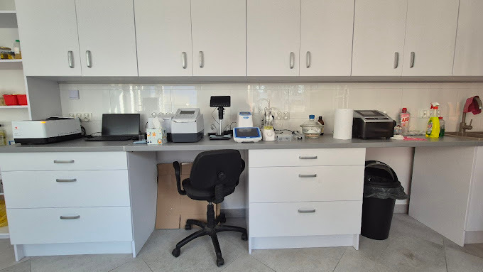Laboratorium z białymi szafkami, sprzętem analitycznym i biurowym krzesłem. Widoczny blat z różnymi urządzeniami i akcesoriami laboratoryjnymi.