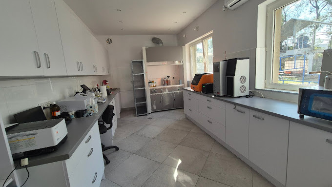 Jasne laboratorium z białymi szafkami, sprzętem analitycznym i oknem z widokiem na zewnątrz. Widoczny wyciąg laboratoryjny i różnorodna aparatura.