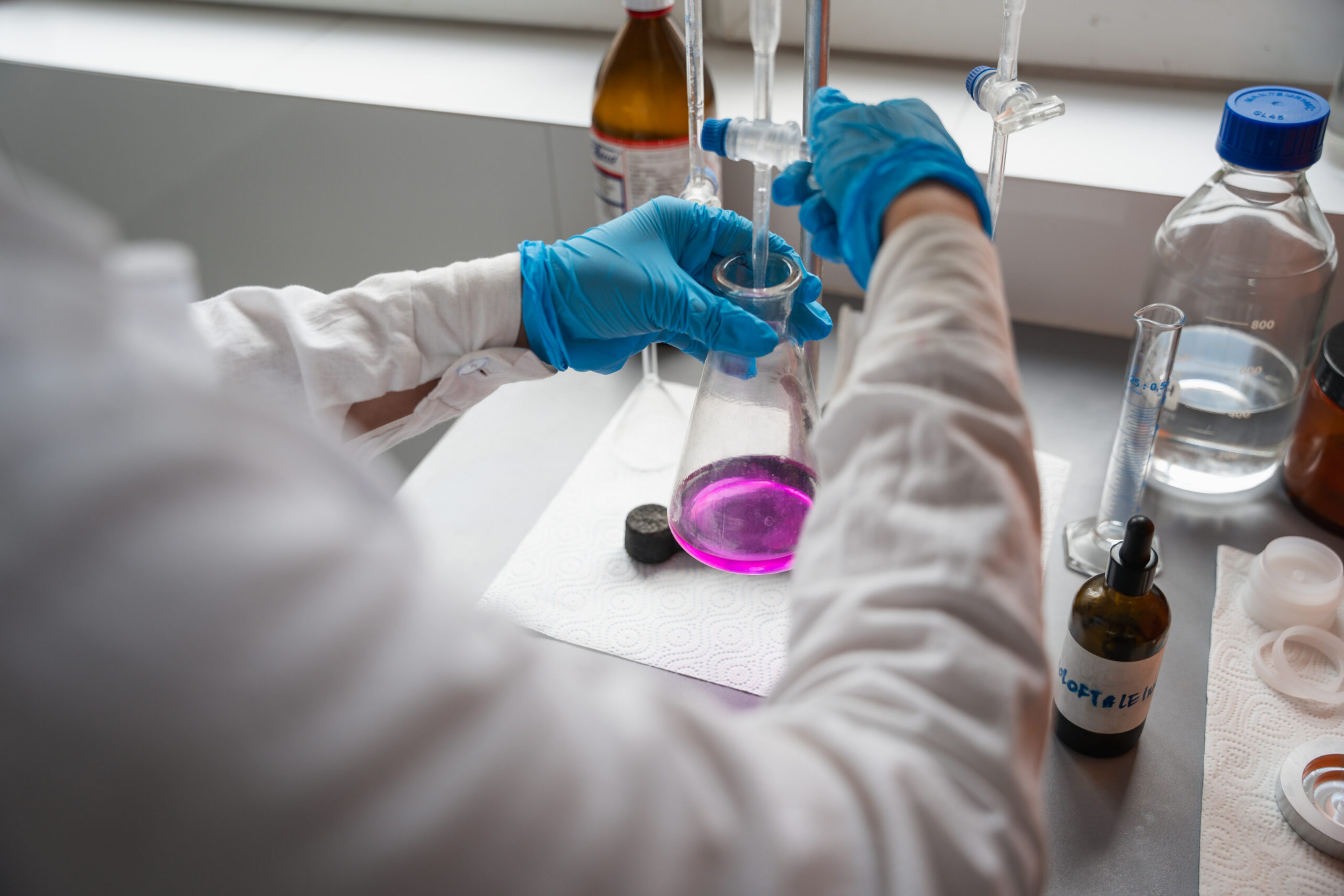 Laboratorium: osoba w rękawicach miesza probówkę z fioletowym płynem, otoczona szkłem laboratoryjnym i odczynnikami na czystym blacie.