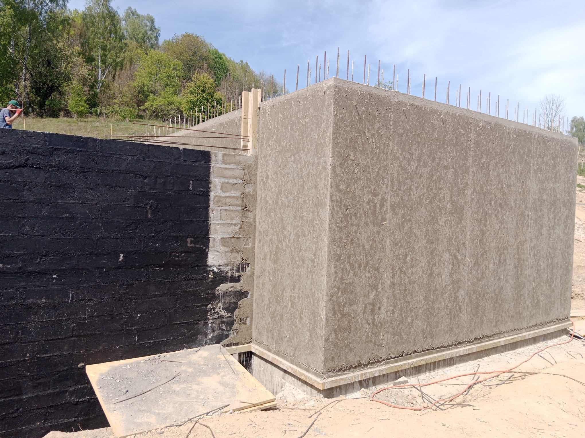 Betonowa konstrukcja w trakcie budowy z widocznym zbrojeniem i czarną izolacją przeciwwilgociową, na tle zieleni i błękitnego nieba.