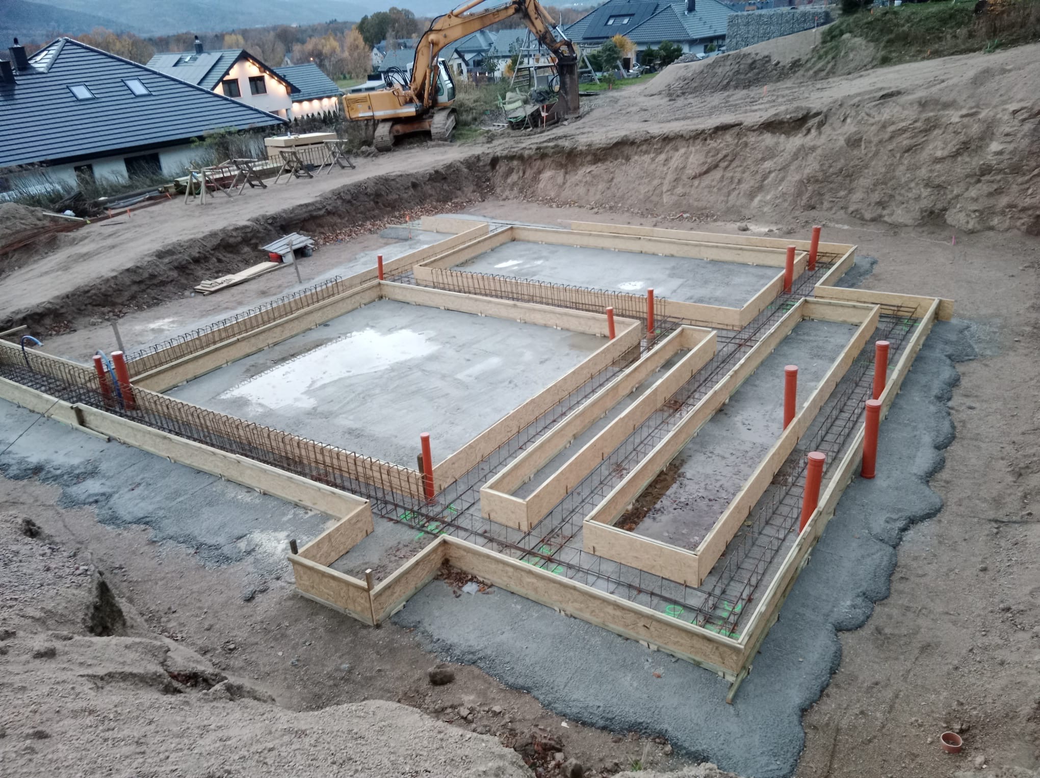 Zalane fundamenty domu z deskowaniem i zbrojeniem, widok z góry. Na drugim planie koparka i zabudowa jednorodzinna w jesiennej scenerii.