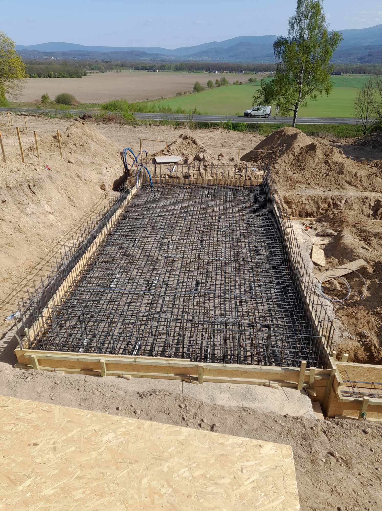 Zbrojenie fundamentów pod budynek, widok z góry. Stalowe pręty tworzą siatkę w drewnianym szalunku. W tle krajobraz z drogą i drzewem.