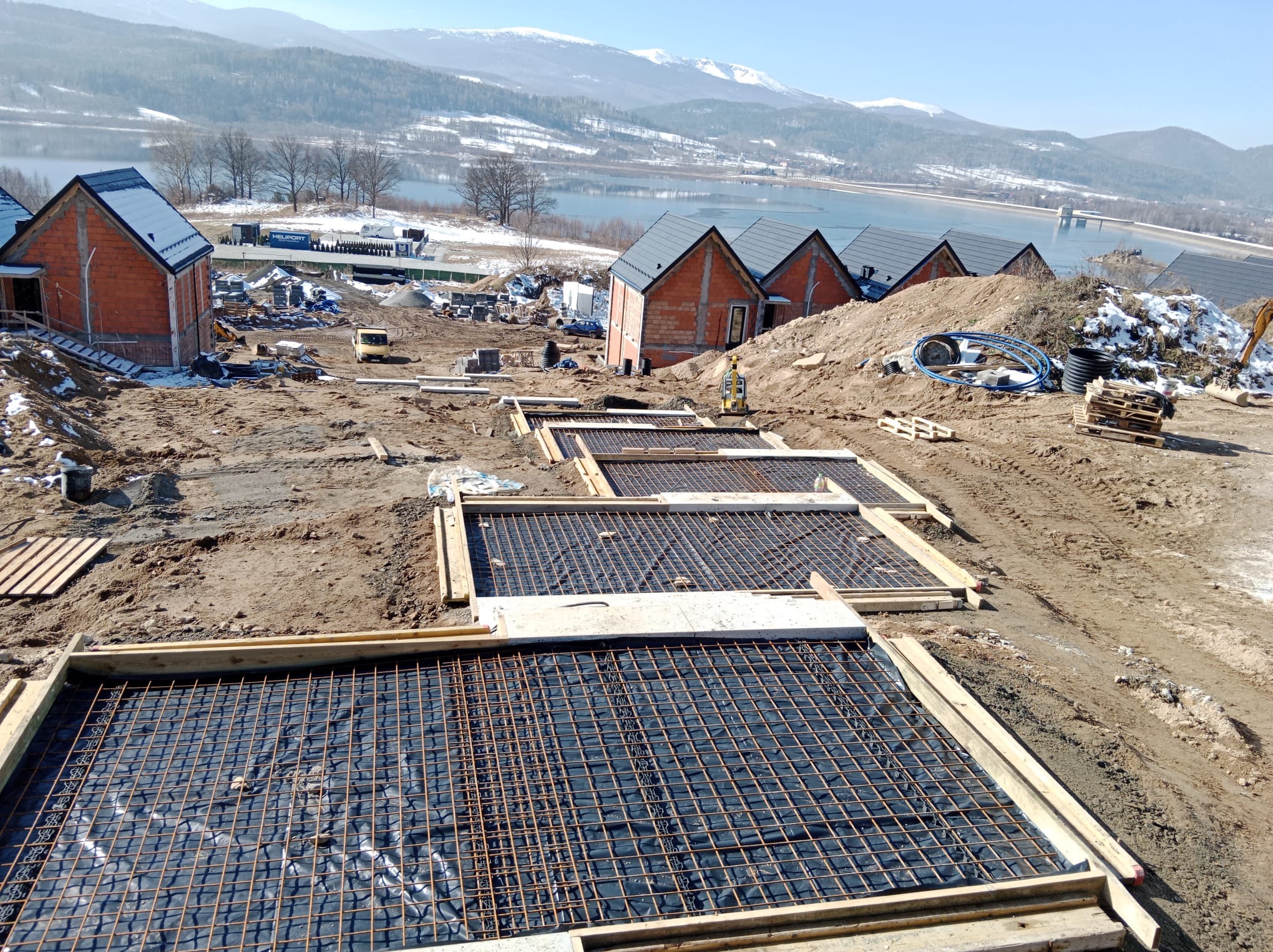 Zbrojenie fundamentów pod budowę domów jednorodzinnych z widokiem na jezioro i góry w tle. Widoczne szalunki i siatka zbrojeniowa.