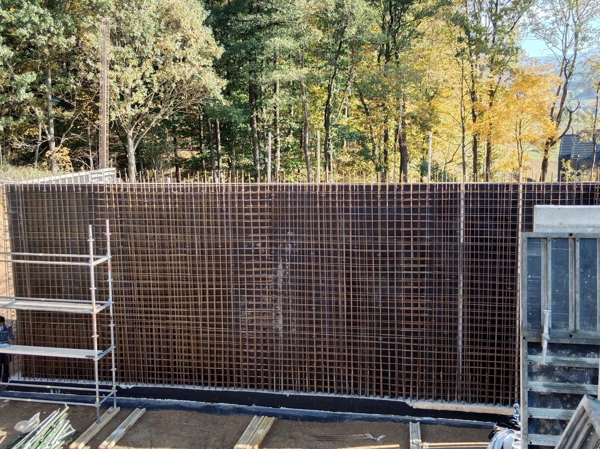 Zbrojenie ściany żelbetowej na tle drzew w jesiennej scenerii. Widoczne rusztowanie i szalunki. Prace zbrojarskie w toku, przygotowanie do betonowania.