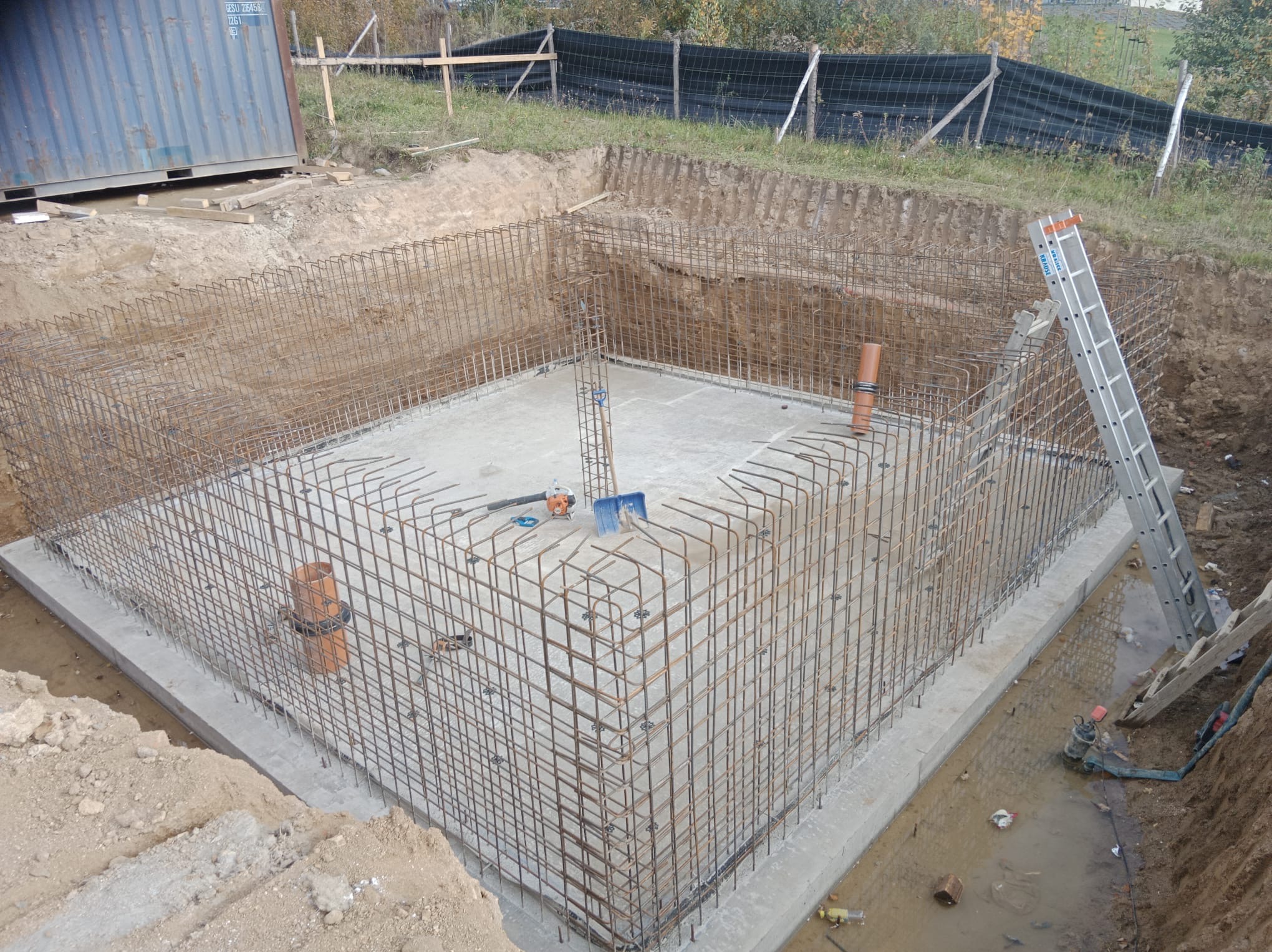 Zbrojenie fundamentu domu jednorodzinnego w wykopie, widok z góry. Stalowe pręty tworzą szkielet, widoczne narzędzia i drabina. Etap budowy przed zalaniem betonem.