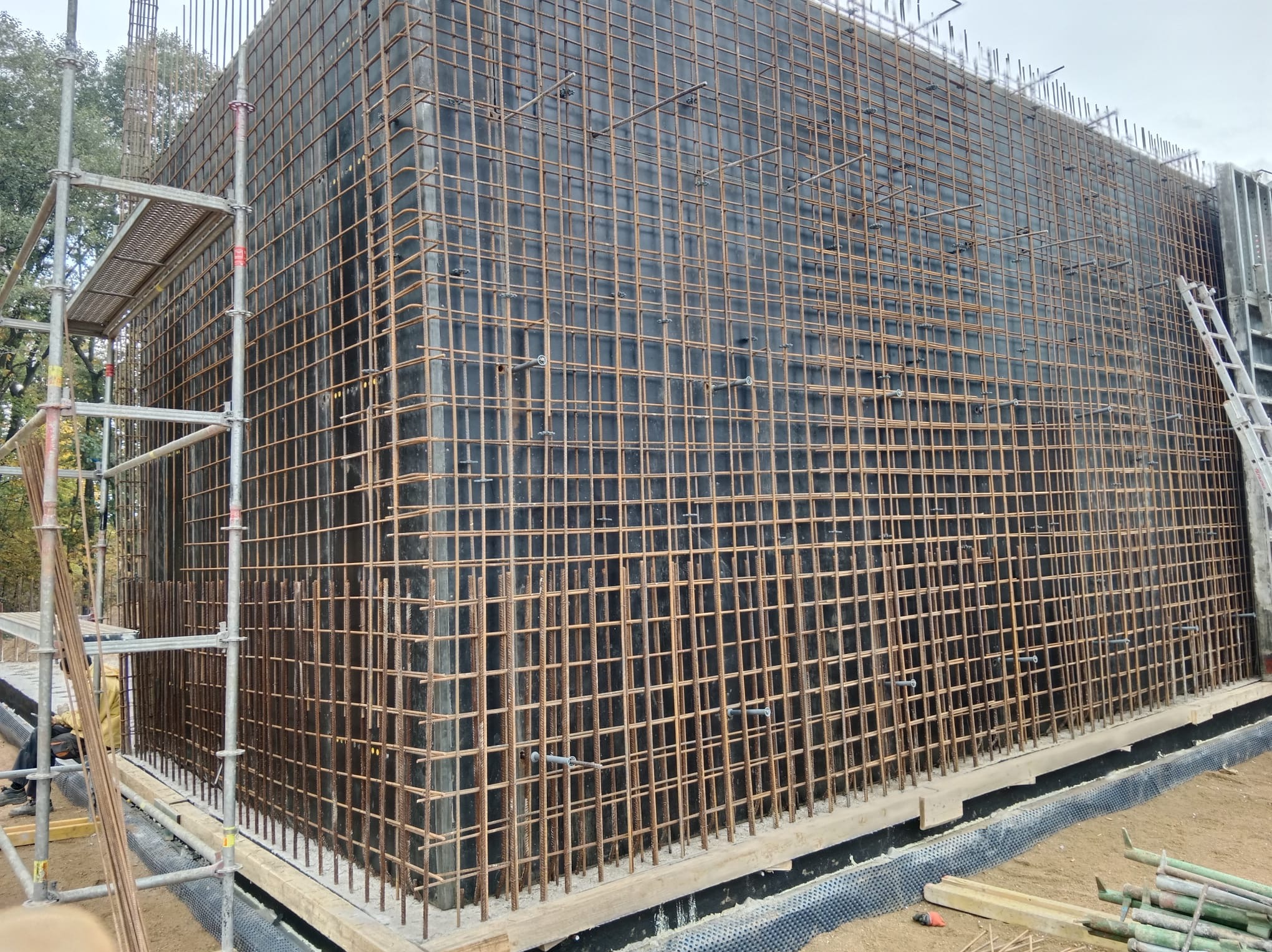 Zbrojenie pionowej ściany szalunkowej z widocznymi prętami zbrojeniowymi, rusztowaniem i elementami konstrukcyjnymi na placu budowy. Widok z dołu, oświetlenie dzienne.