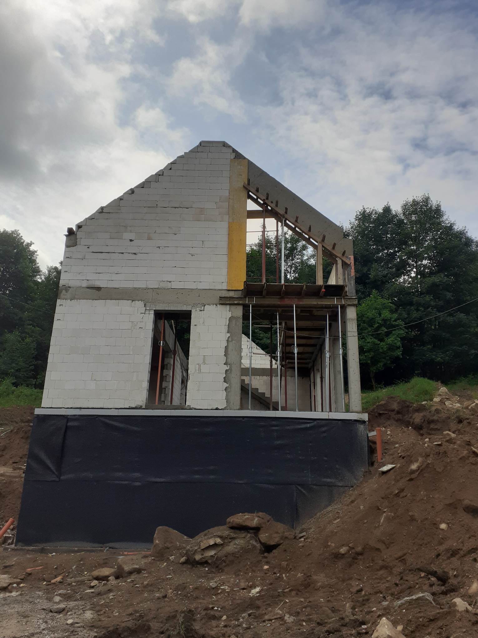 Niedokończony dom z białego pustaka, widoczne zbrojenia i szalunki, czarna izolacja fundamentów, ziemia i drzewa w tle, pochmurne niebo.