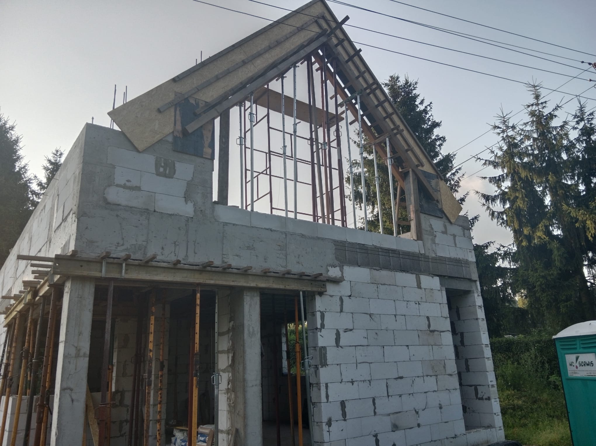 Budowa domu w trakcie: ściany z betonu komórkowego, drewniana konstrukcja dachu z metalowymi podporami. Widoczne drzewa w tle i przenośna toaleta.