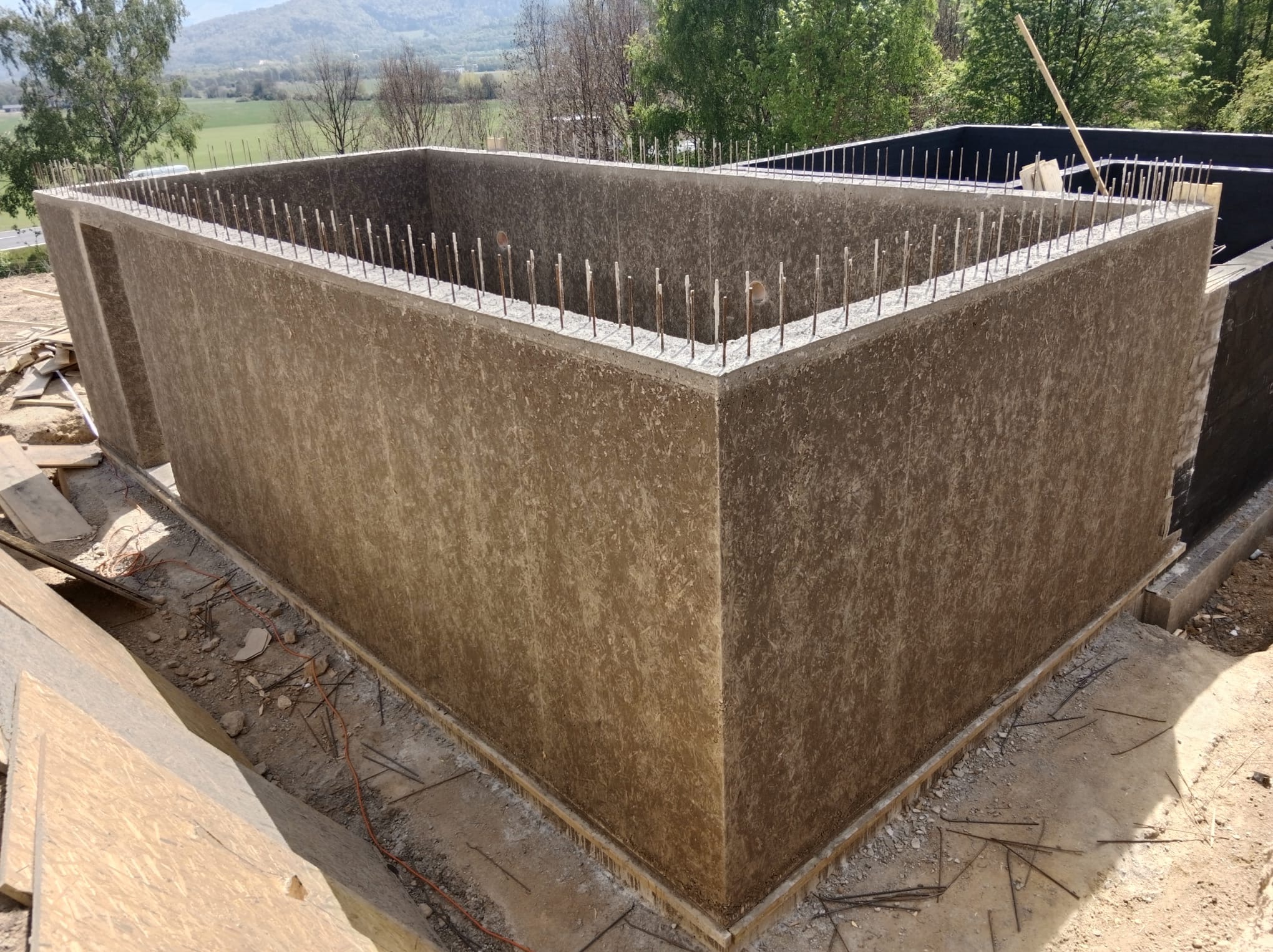 Surowa bryła budynku w trakcie budowy, z widocznymi prętami zbrojeniowymi na górze ścian. Tło stanowią drzewa i wzgórza.