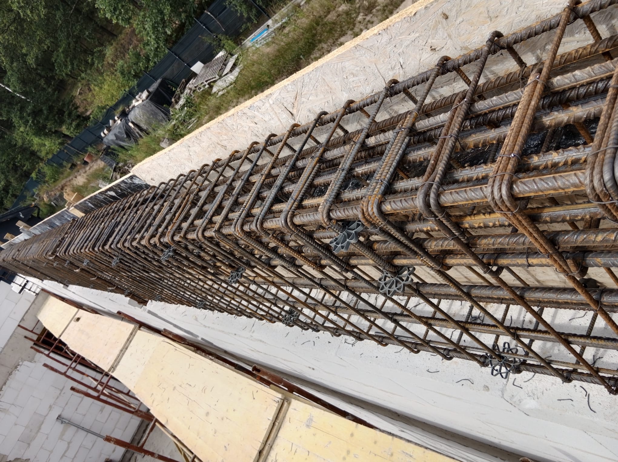Zbrojenie belki stropowej z dystansami, widok z góry, na tle szalunku z desek i fragmentu ściany z betonu komórkowego. Detal konstrukcji żelbetowej.