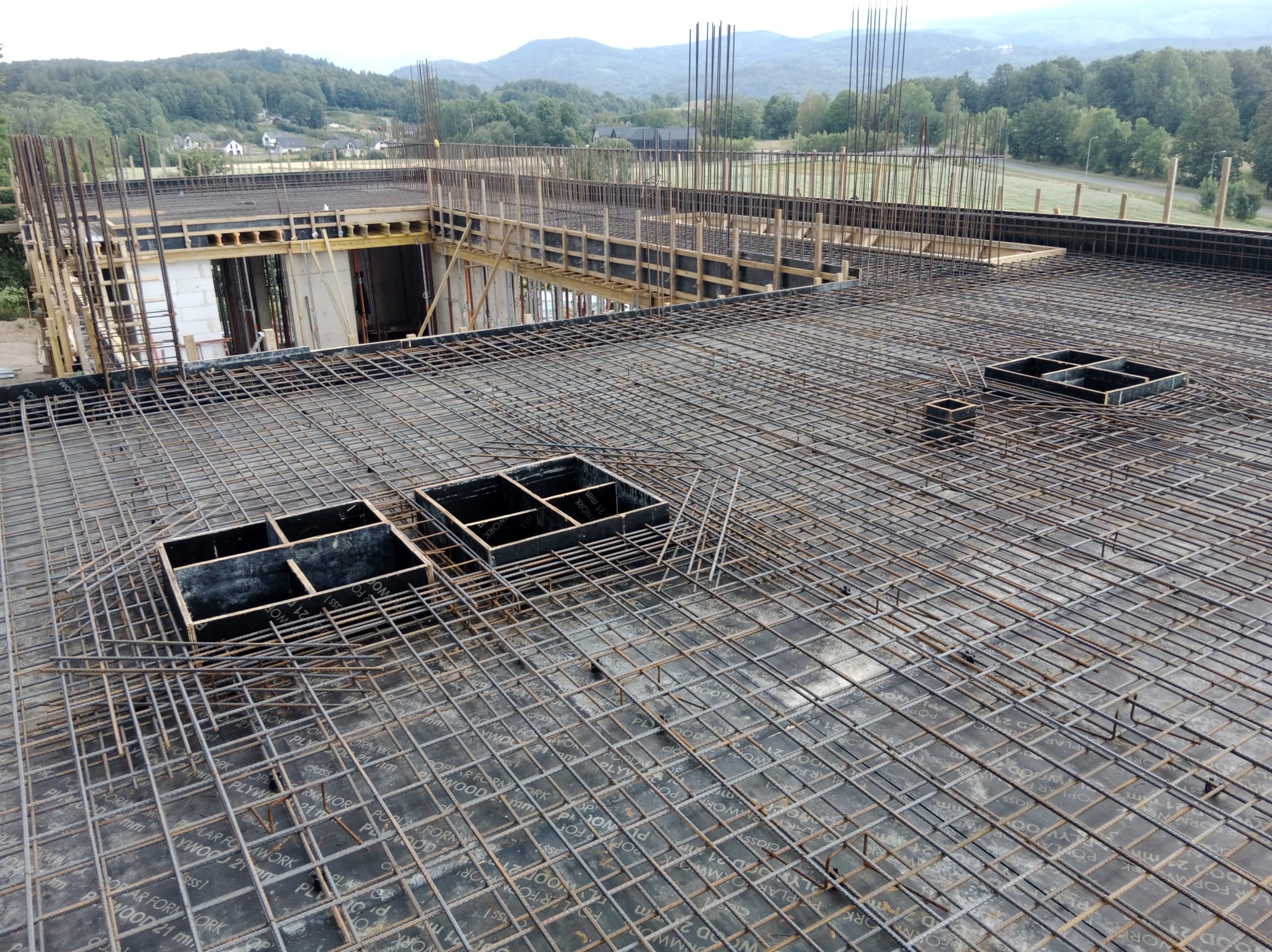 Zbrojenie stropu budynku w trakcie budowy, widoczne szalunki i wystające pręty. W tle zalesione wzgórza.