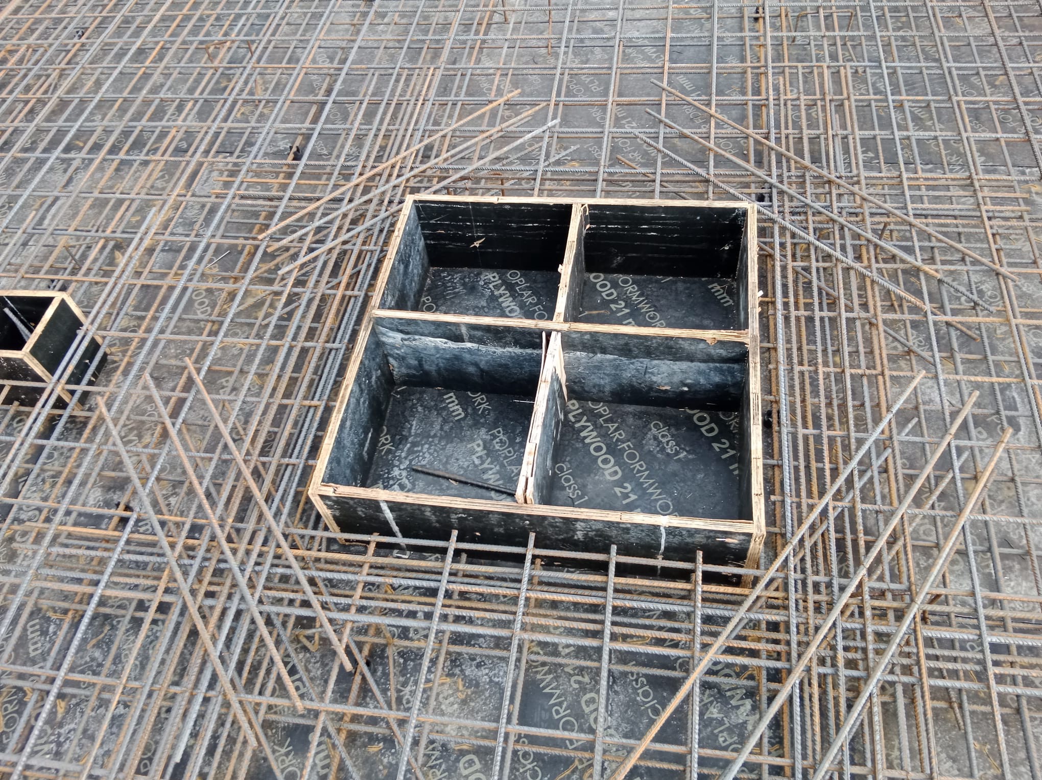 Zbrojenie płyty żelbetowej z drewnianym szalunkiem na otwory, widok z góry. Siatka z prętów stalowych tworzy regularną strukturę. Szalunek z czarnej sklejki.