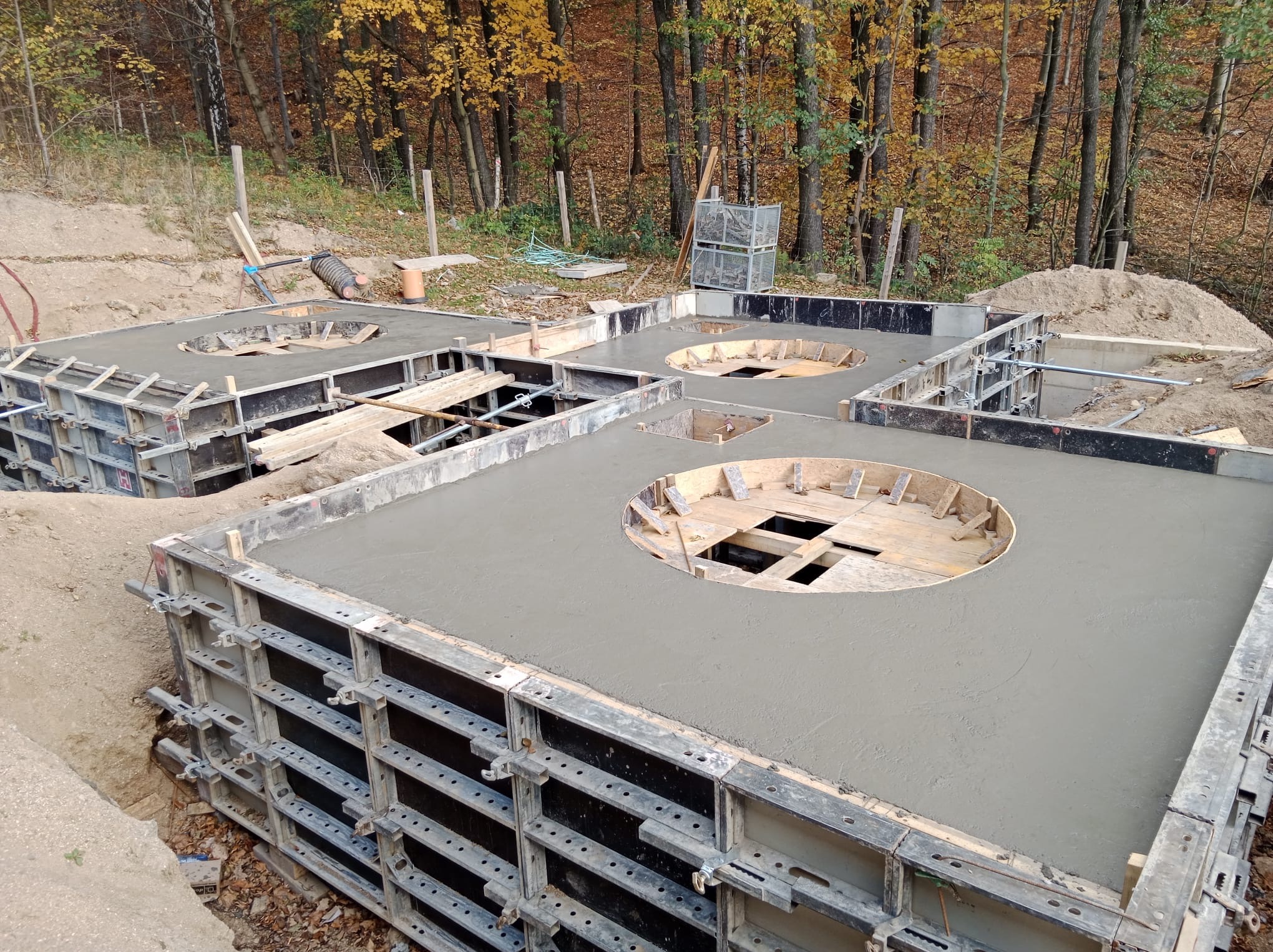 Zalewanie betonem fundamentów z okrągłymi otworami w szalunkach na tle jesiennego lasu. Widoczne drewniane elementy konstrukcji i metalowe szalunki.