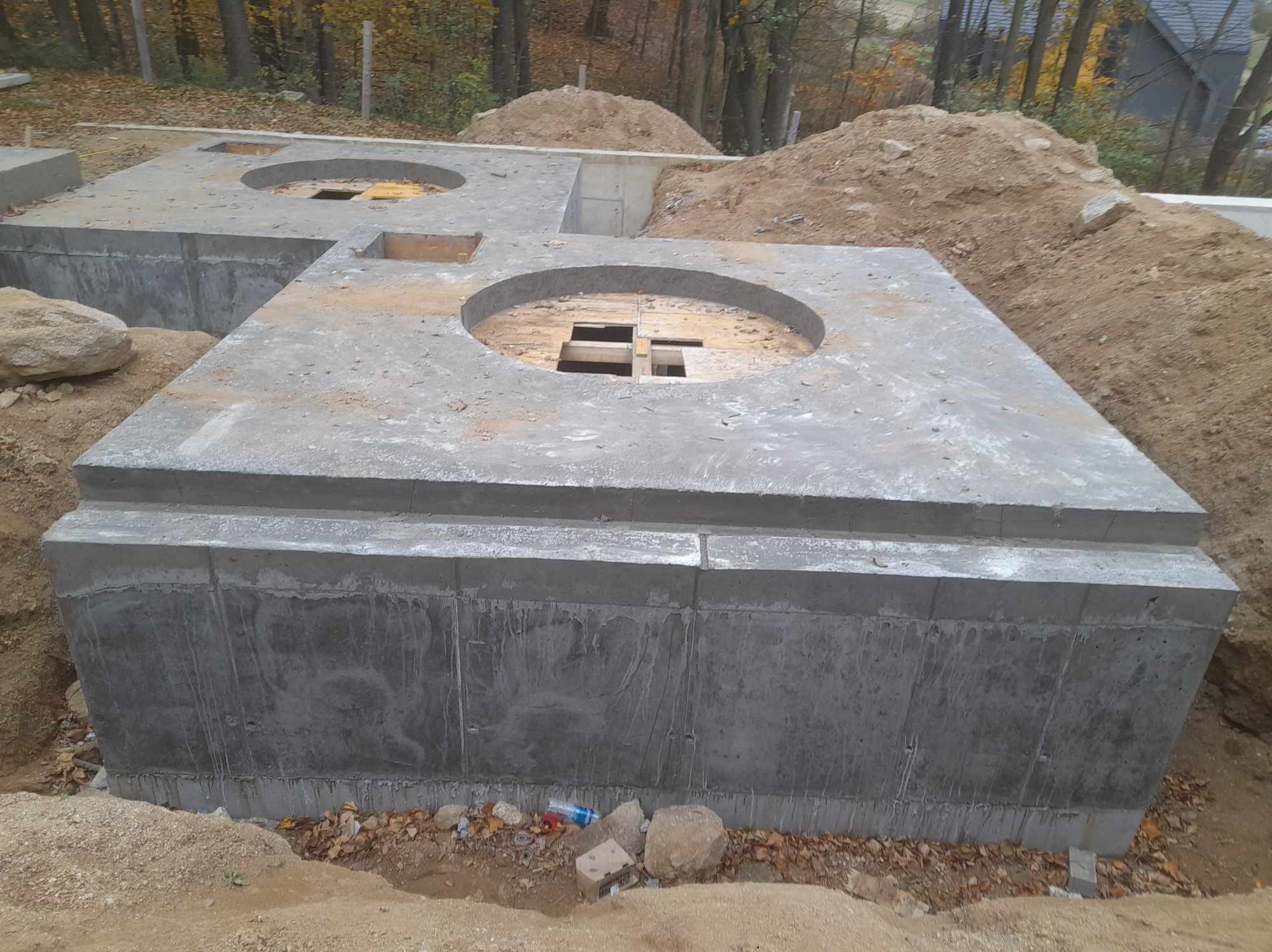 Betonowe fundamenty z okrągłymi otworami w ziemi, przygotowane pod budowę domu. Widoczne ślady po szalunkach i wykopana ziemia w tle.