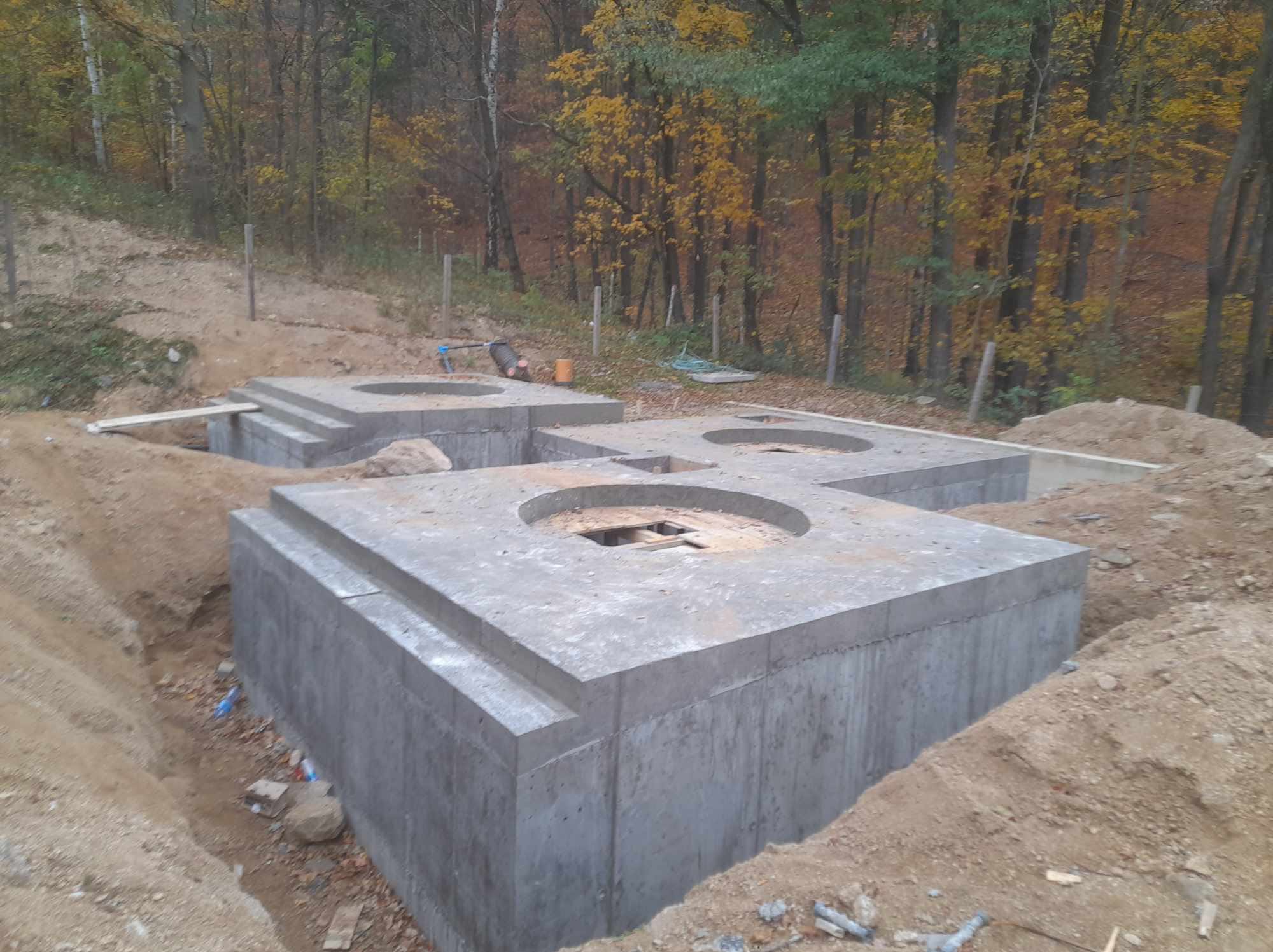 Betonowe fundamenty z okrągłymi otworami na tle jesiennego lasu. Widoczne ślady wykopów i przygotowania terenu pod budowę. Surowy beton w naturalnym otoczeniu.